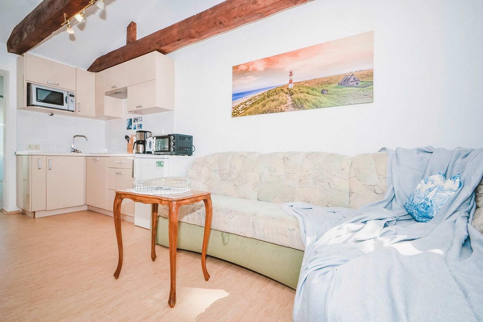 Living room with sofa, kitchenette, and wooden ceiling beams. Wall art shows lighthouse.