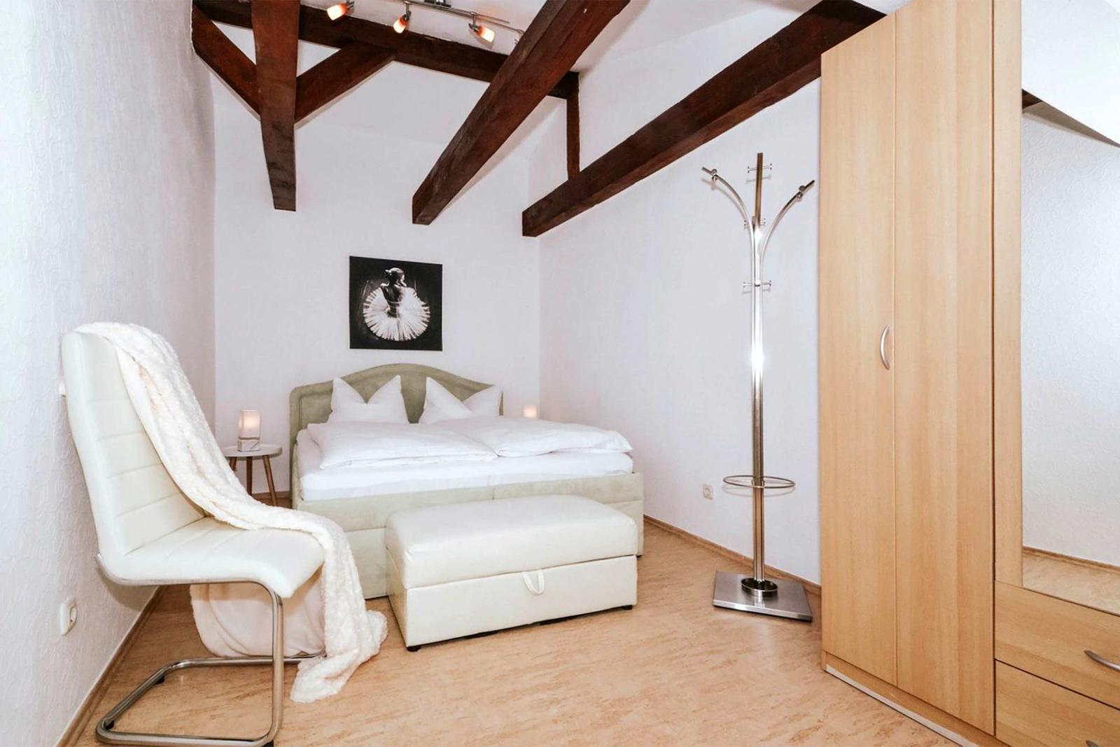 Bedroom with bed, chair, coat stand, and wardrobe under wooden beam ceiling.