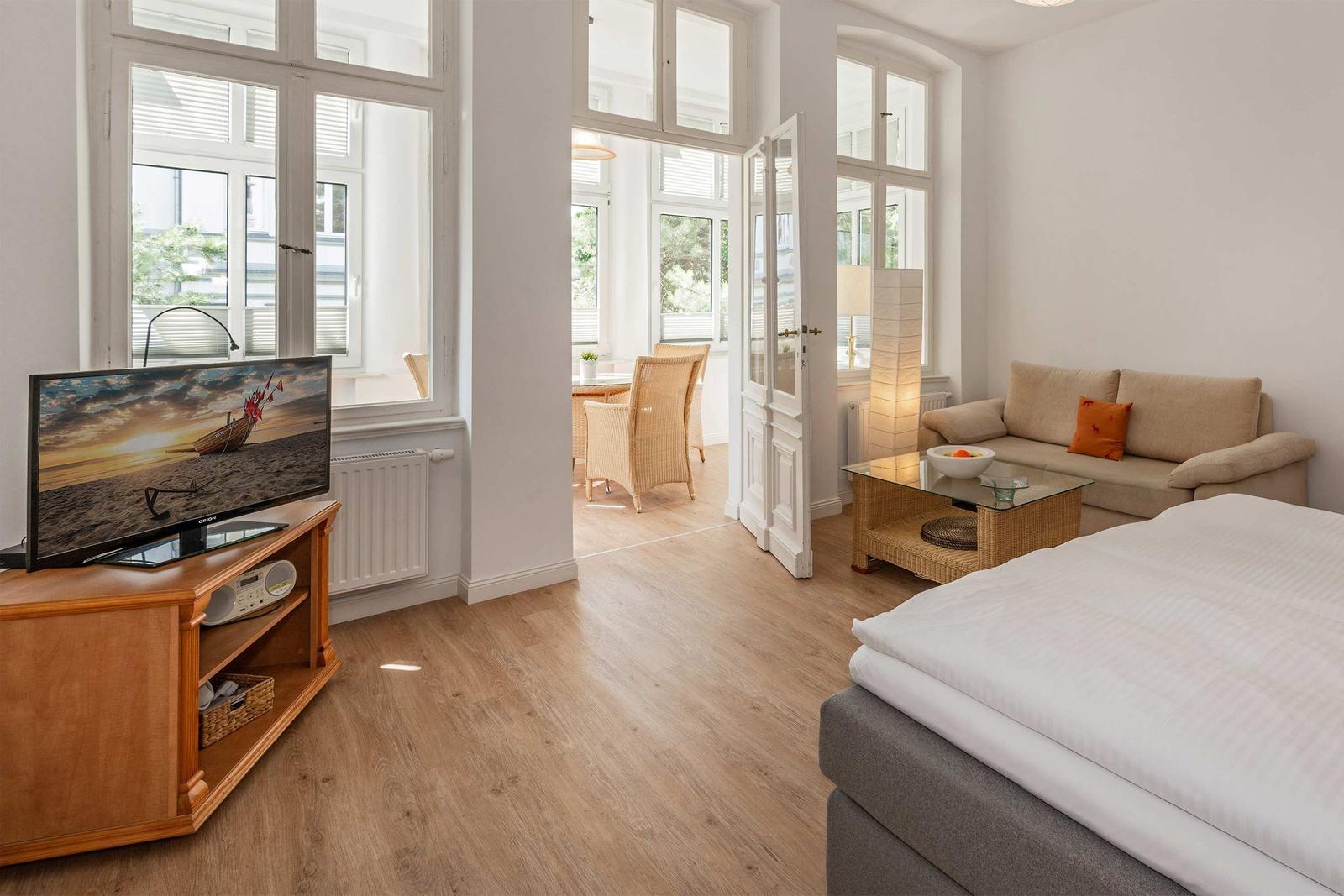 A bedroom with bed, sofa, and TV. Continuous hardwood flooring. Doors lead to a dining area.