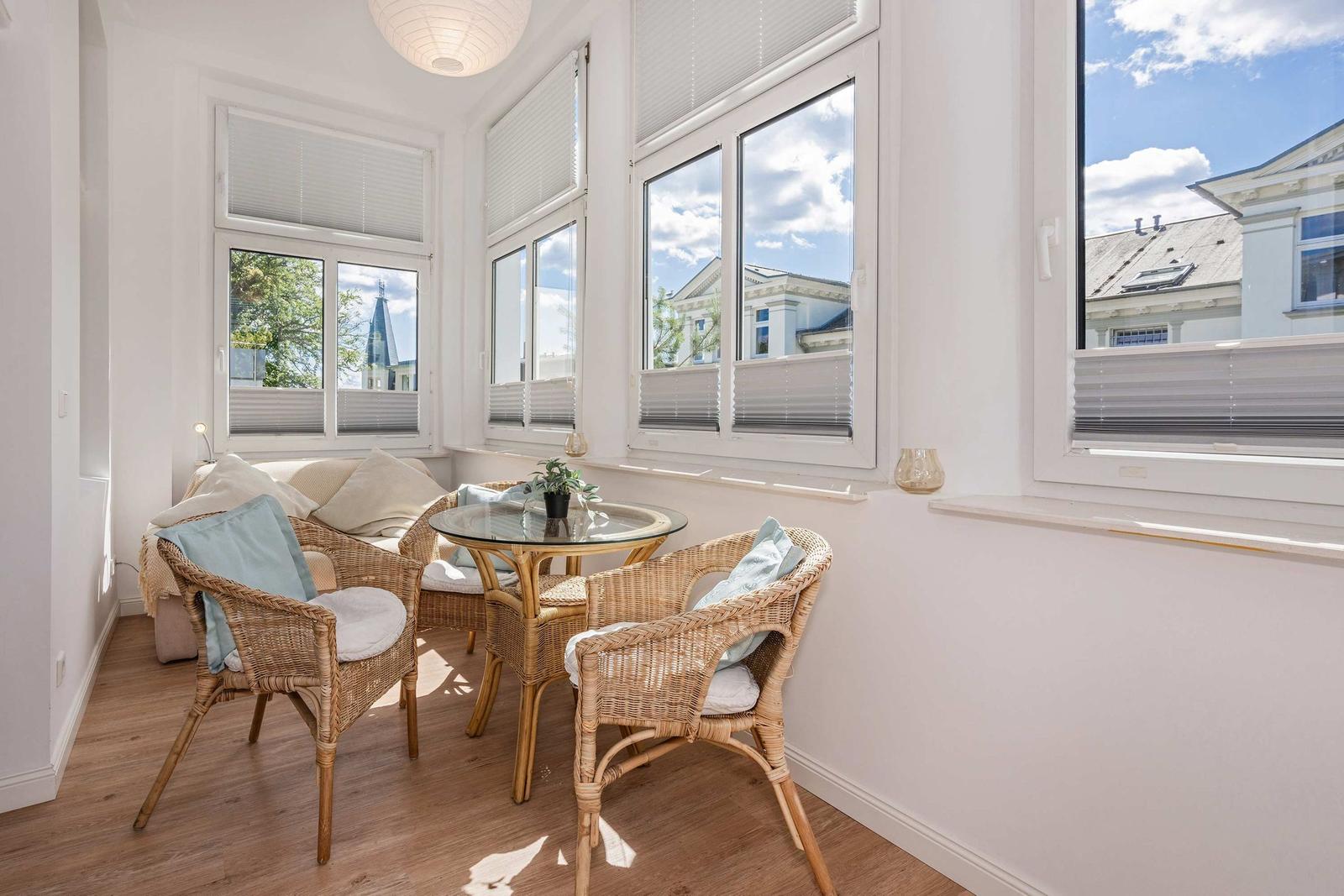 Living area with rattan furniture, glass table, and large windows overlooking buildings.