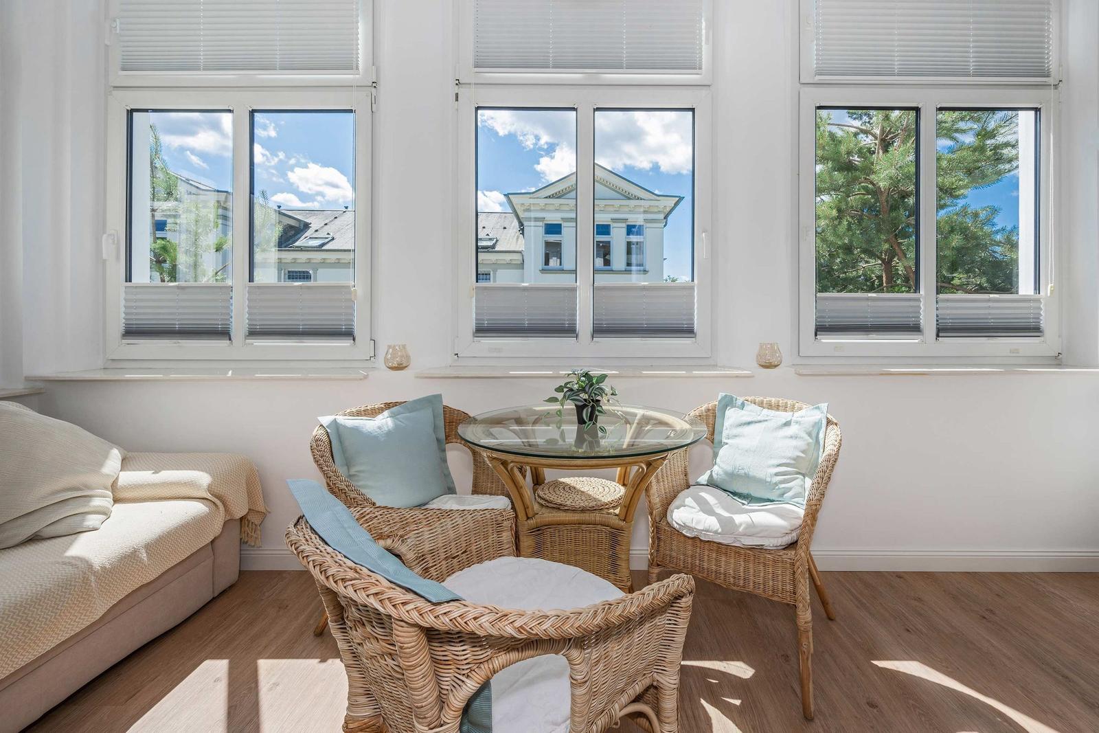 Living room with rattan furniture, glass table, and view of buildings and trees.