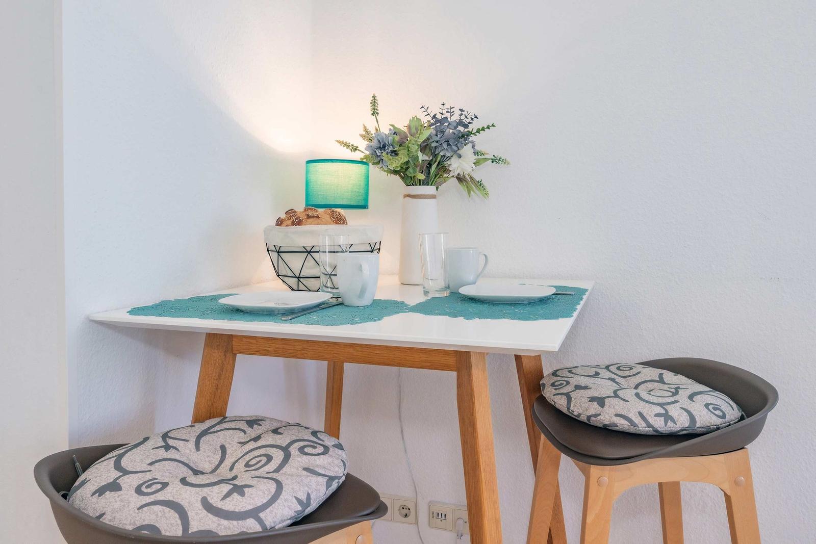 Esstisch mit zwei Hockern, Tischdecke, Tassen und Blumenarrangement.