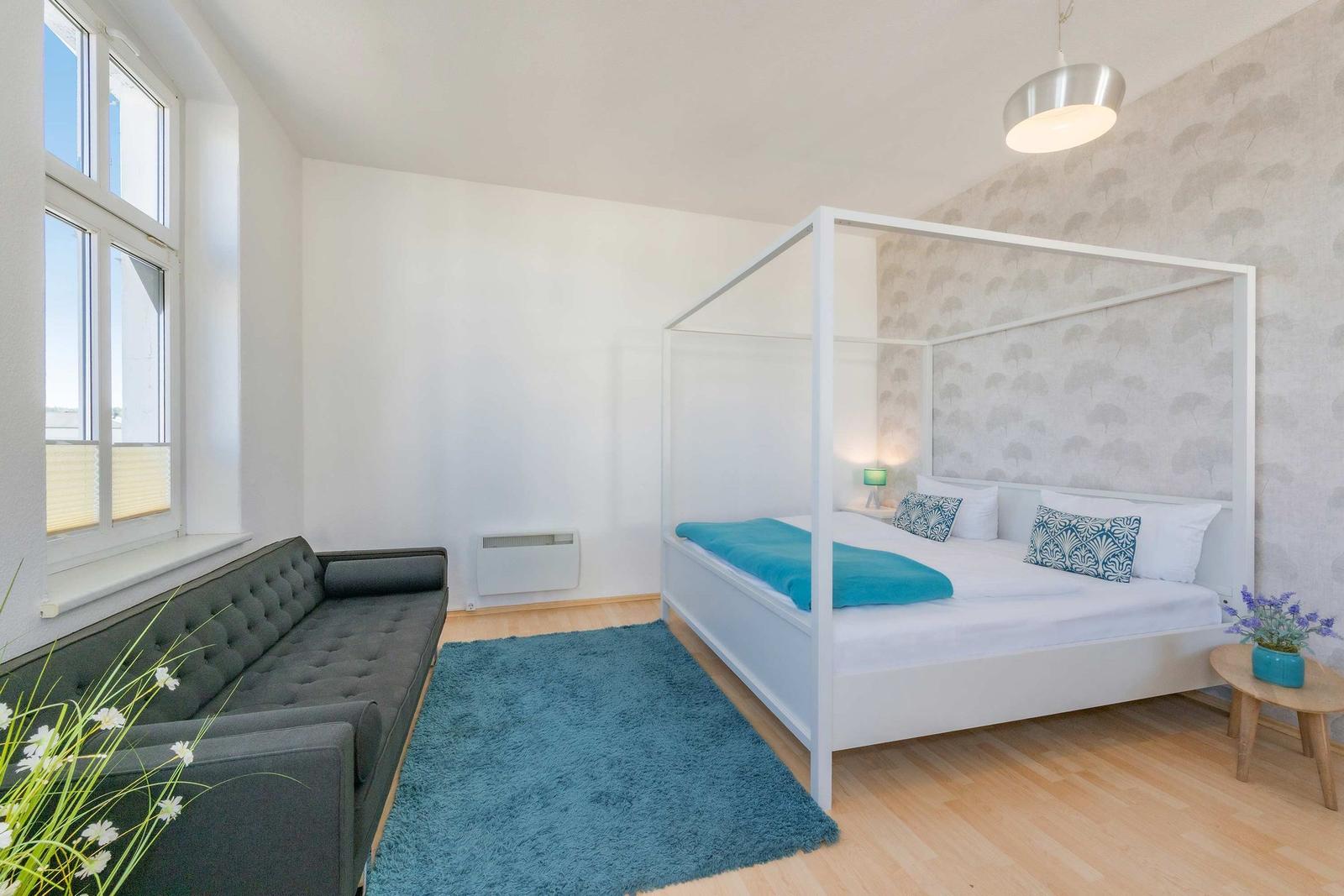 Schlafzimmer mit weißem Bett, grauem Sofa und blauem Teppich. Fenster und Holzboden.