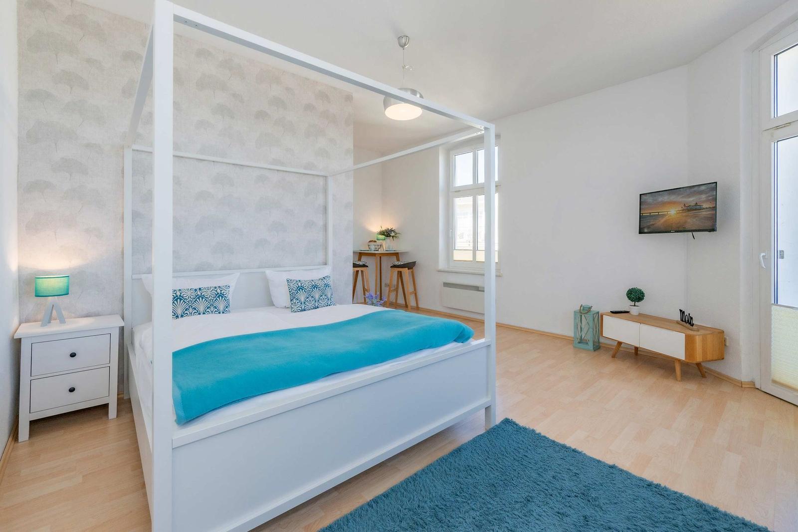 Schlafzimmer mit weißem Bett, blauer Decke und Holzboden. Fernseher an der Wand.