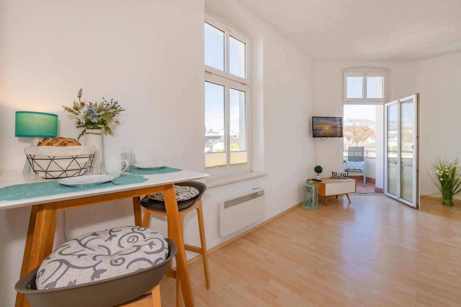 Wohnraum mit Esstisch, Fenstern und Balkontür. Holzboden und weiße Wände.