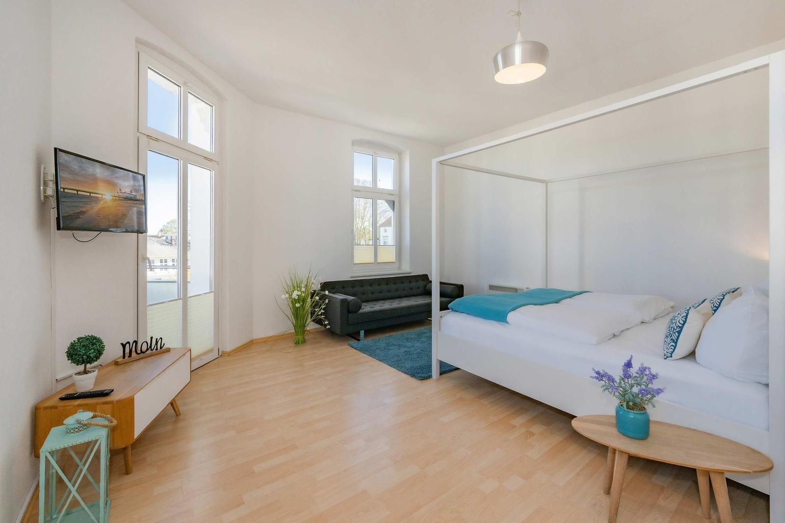Schlafzimmer mit Bett, Sofa, TV und Holzboden. Fenster mit Blick nach draußen.