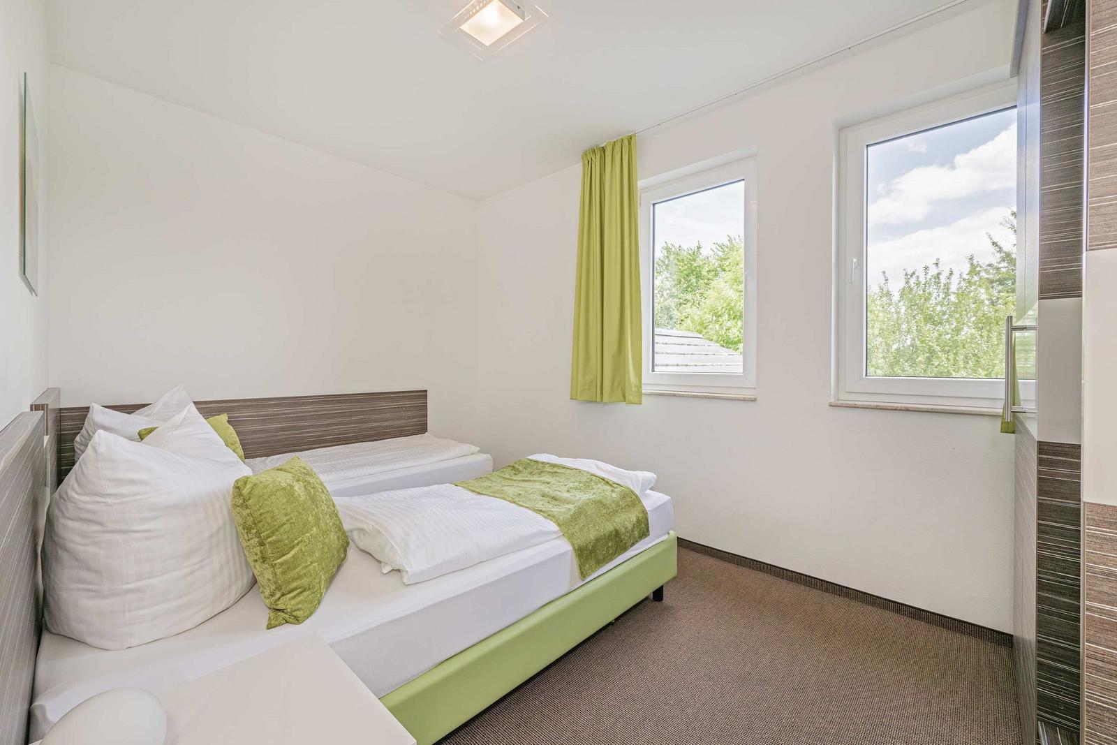 Zimmer mit Doppelbett, grünen Kissen und Blick auf Bäume durch Fenster.