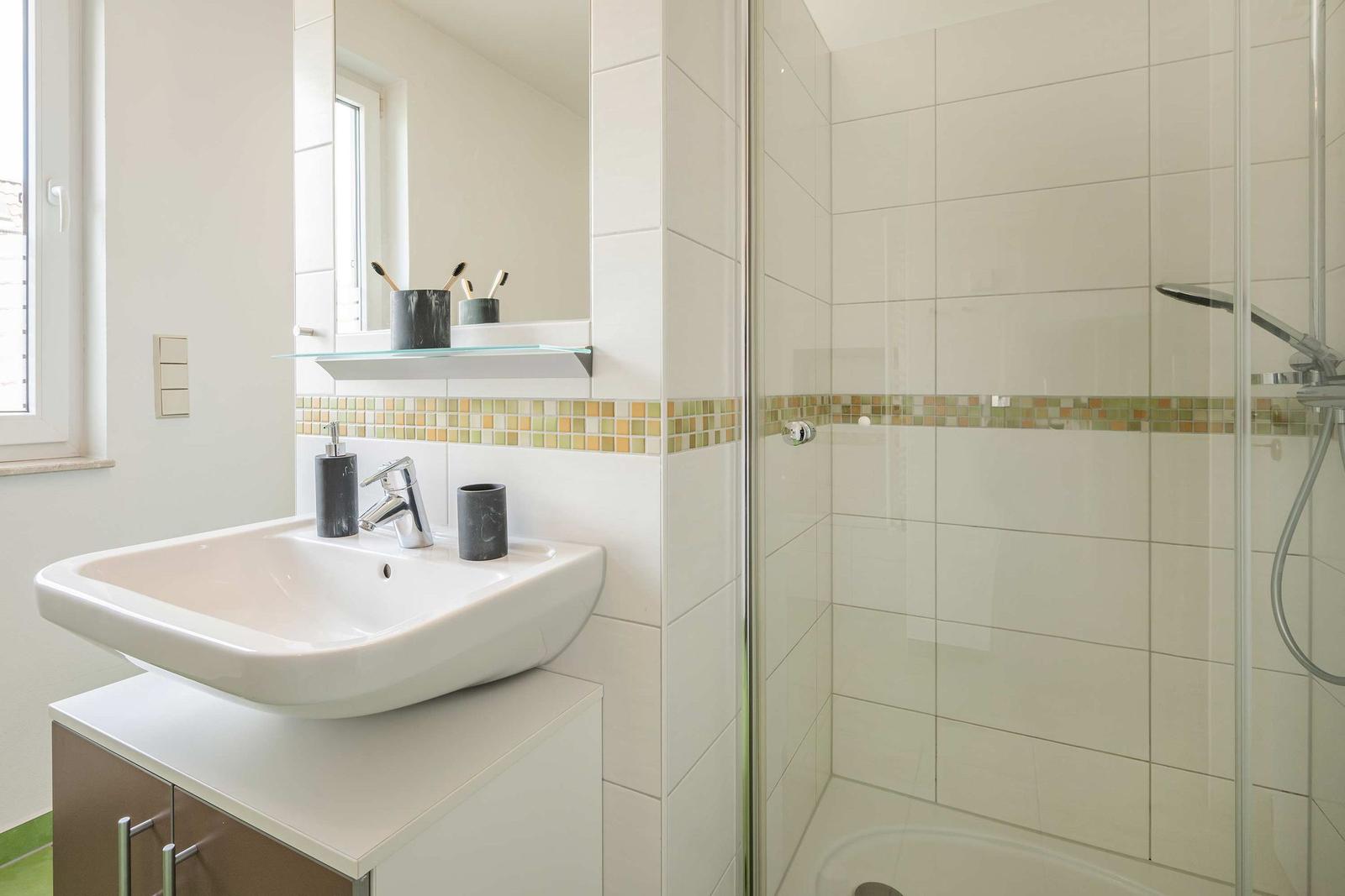 Badezimmer mit Dusche, Waschbecken und Spiegel. Wandfliesen mit Mosaikband.