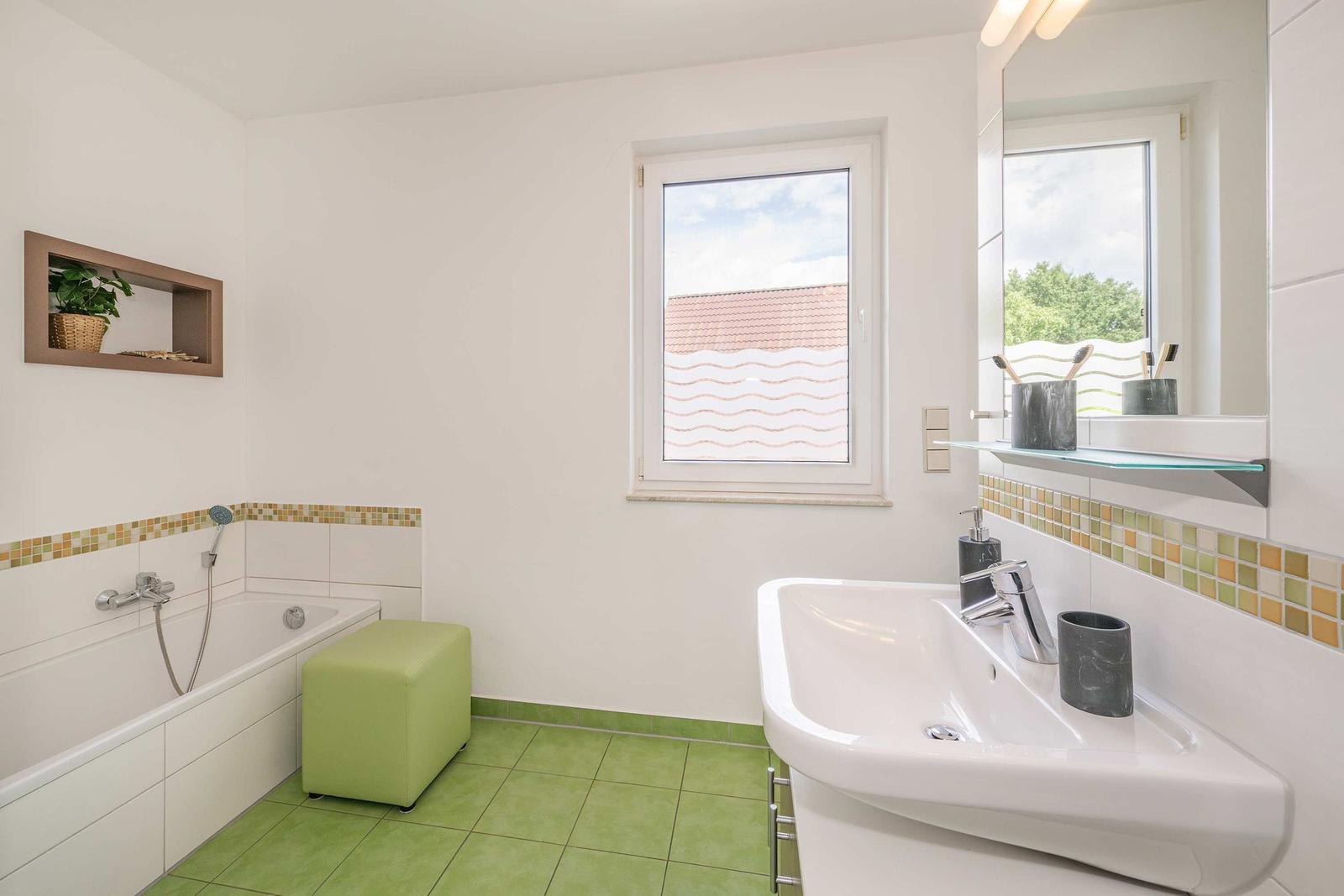 Badezimmer mit Badewanne, Waschbecken und Fenster. Grün gefliest und mit Mosaik verziert.