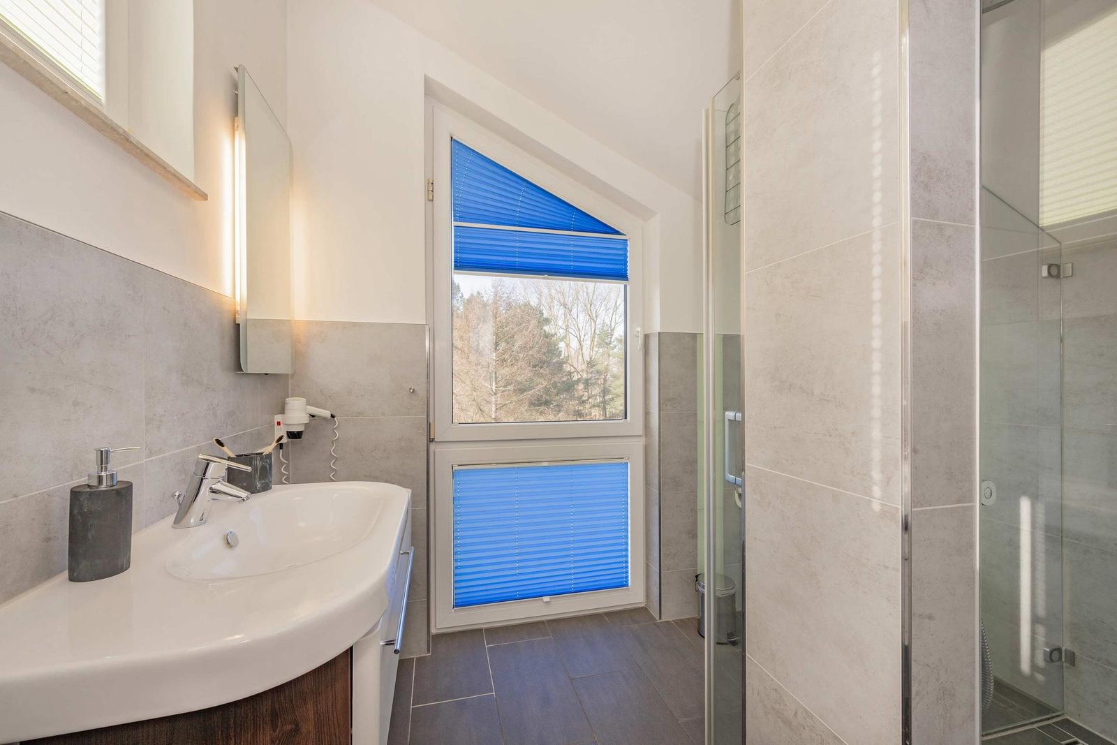 Badezimmer mit Dusche, Waschbecken und Fenster mit blauen Jalousien.