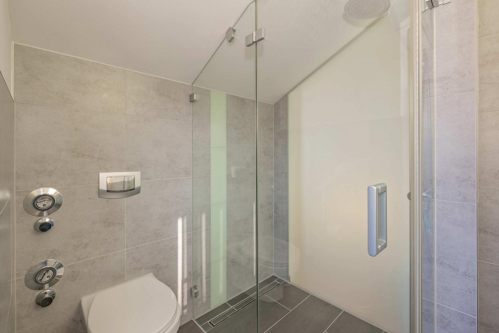 Dusche mit Glaswand, Toilette und Wandarmaturen in modernem Badezimmer.