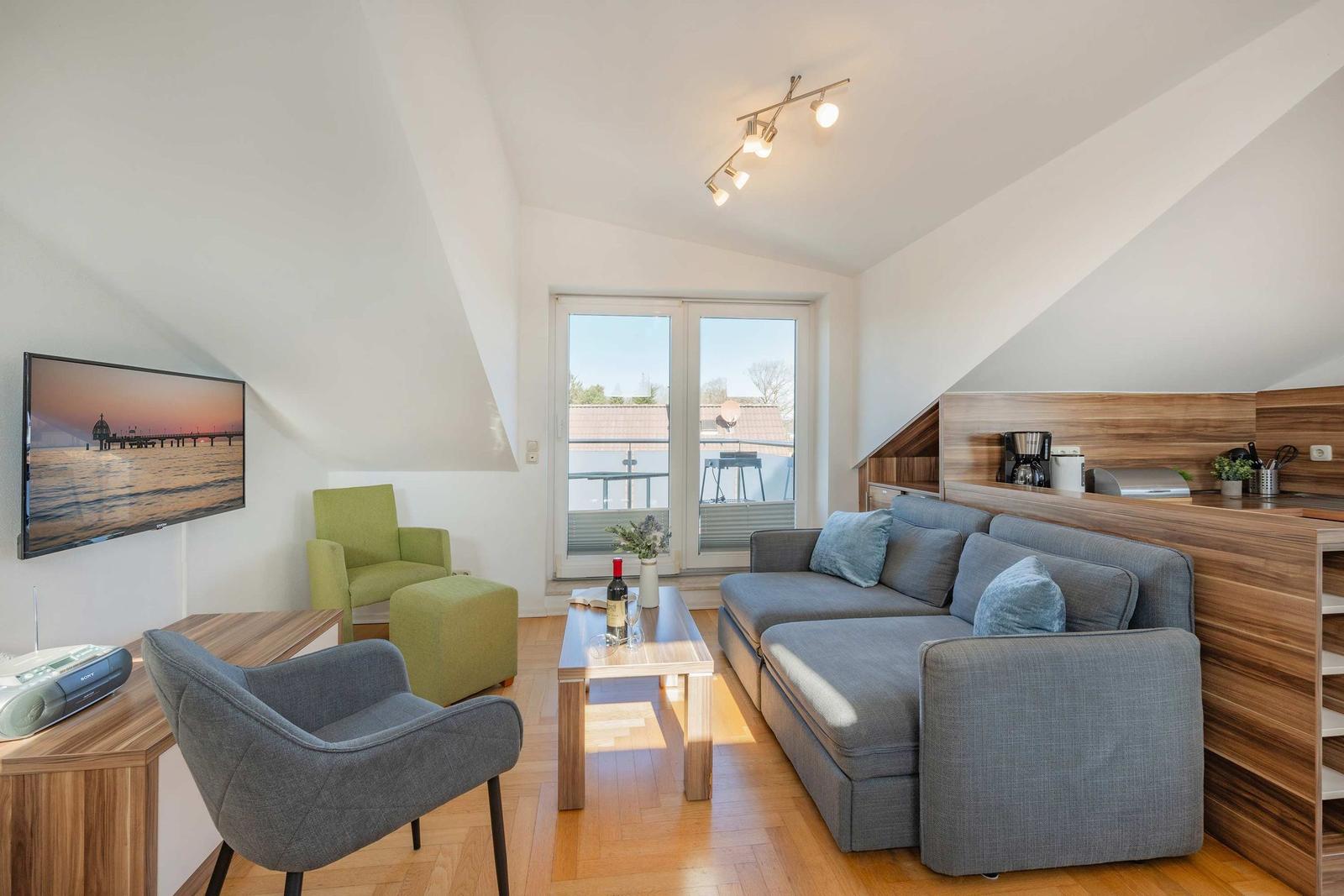 Wohnbereich mit Sofa, Sessel und TV. Balkon und Holzboden sichtbar.