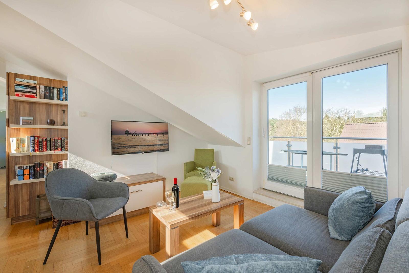 Wohnzimmer mit Sofa, Sessel und Balkon. Fernseher an der Wand. Bücherregal links.