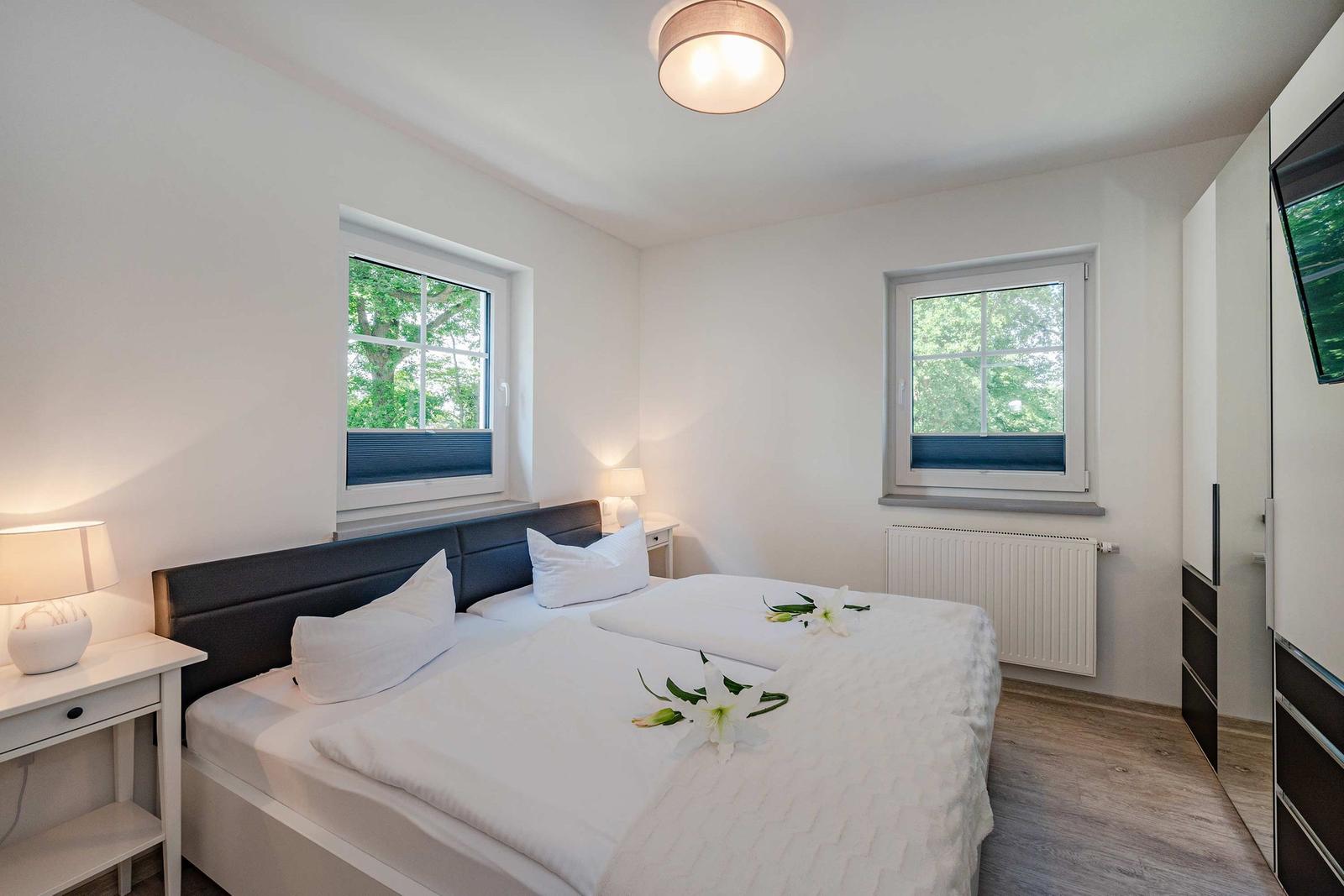 Schlafzimmer mit Doppelbett, zwei Fenstern und Schrank. Bett mit weißen Laken und Blumen.