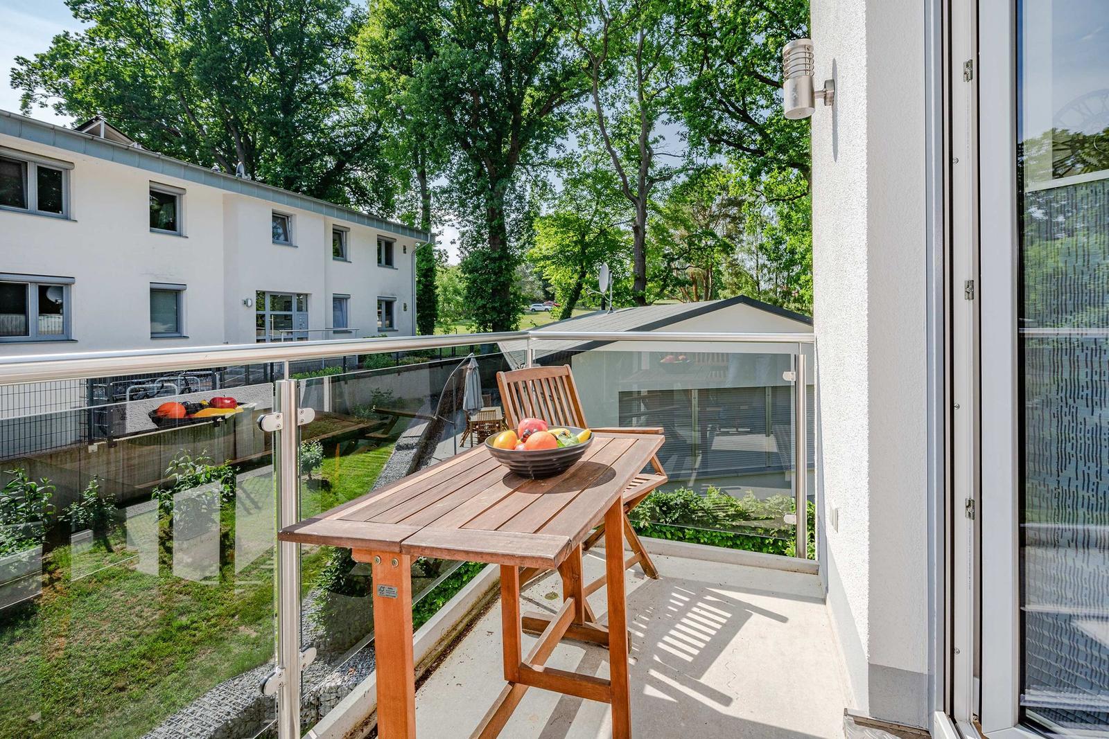 Terrasse mit Holztisch und Stuhl, Obstschale, Glasgeländer und Blick auf Wohnhaus und Bäume.