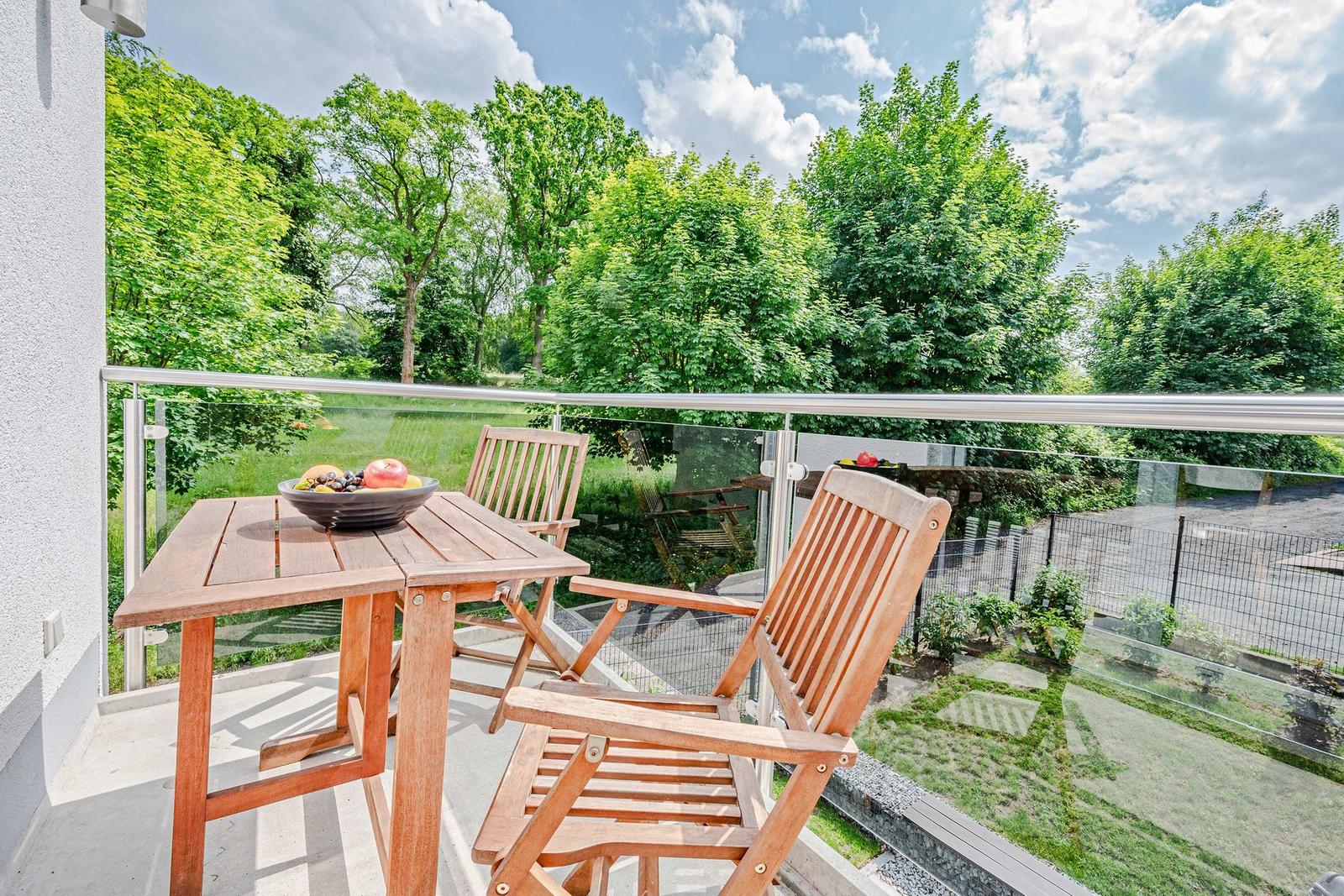 Terrasse mit Holztisch und zwei Stühlen, Blick auf grüne Bäume und einen Garten.