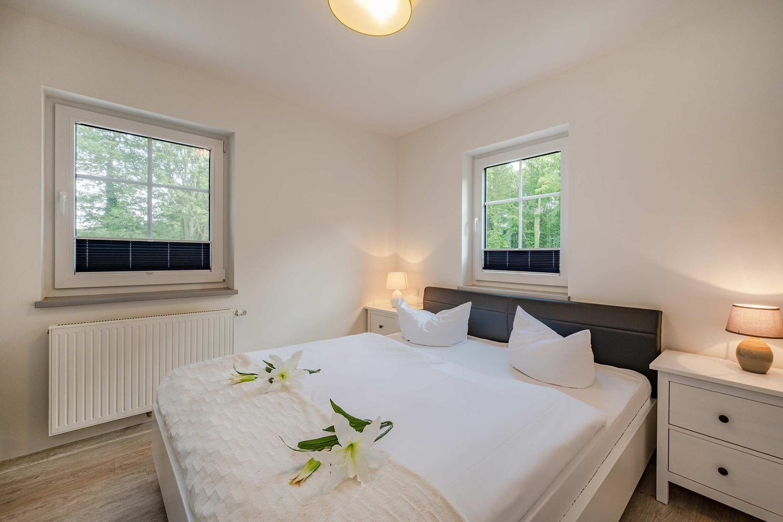 Schlafzimmer mit Doppelbett, zwei Fenstern und Nachttischen mit Lampen.
