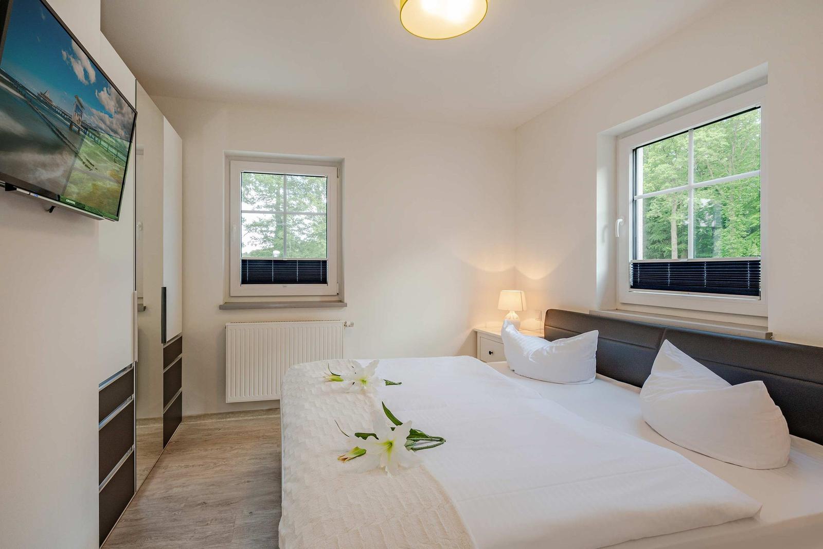 Schlafzimmer mit Bett, Fenstern, TV und Blumen.