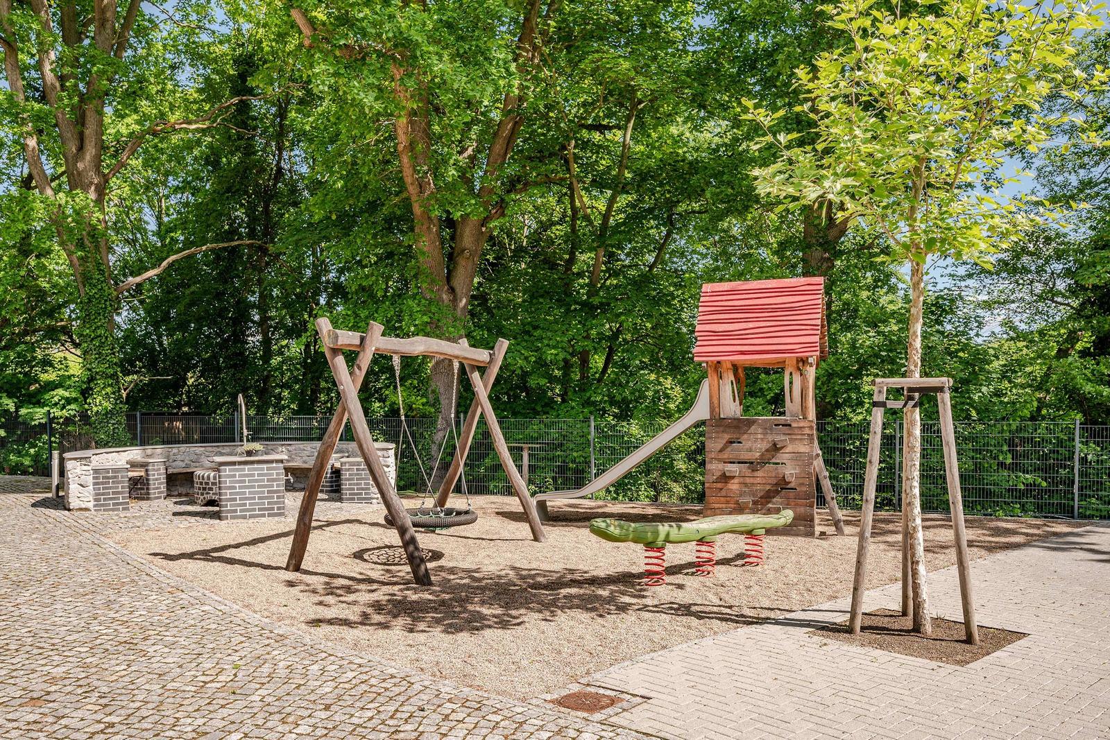 Kinderspielplatz mit Schaukel, Rutsche und Klettergerüst. Umgeben von Bäumen und gepflastertem Weg.