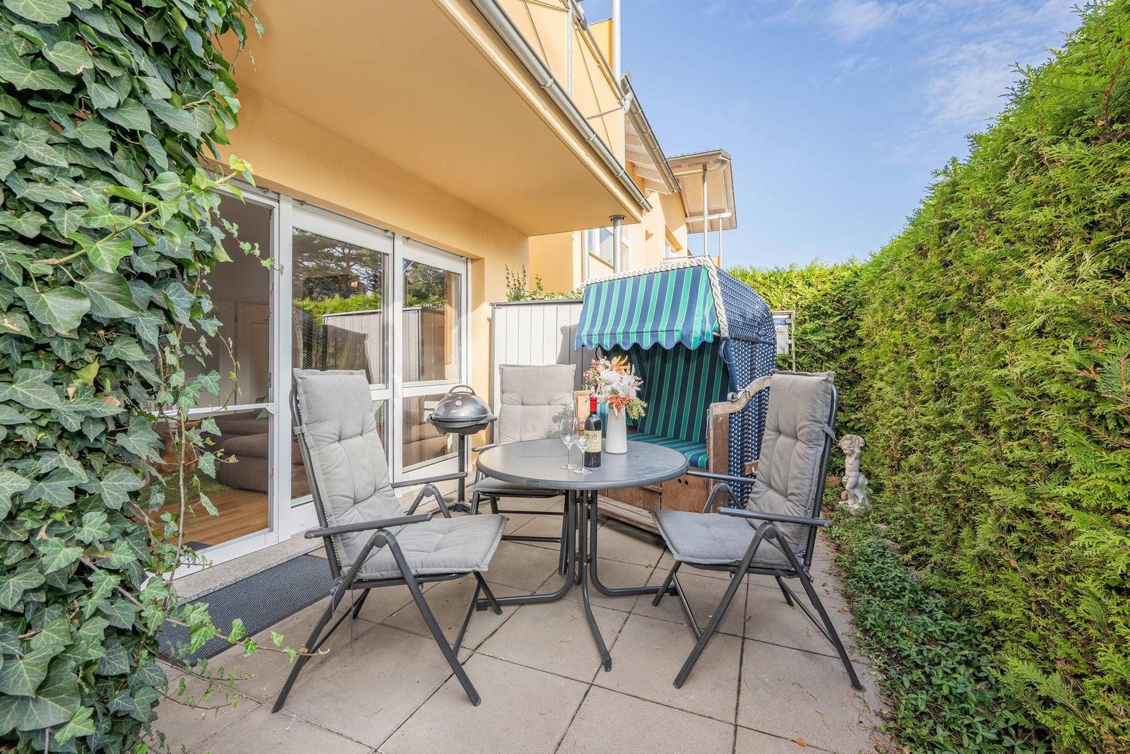 Terrace with table, chairs, and grill. Green hedge and beach chair in background.