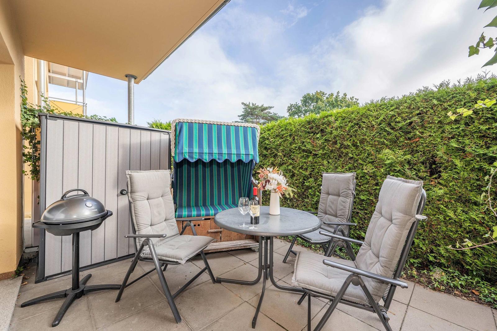 Terrace with table, chairs, grill, and lounge chair behind hedge.
