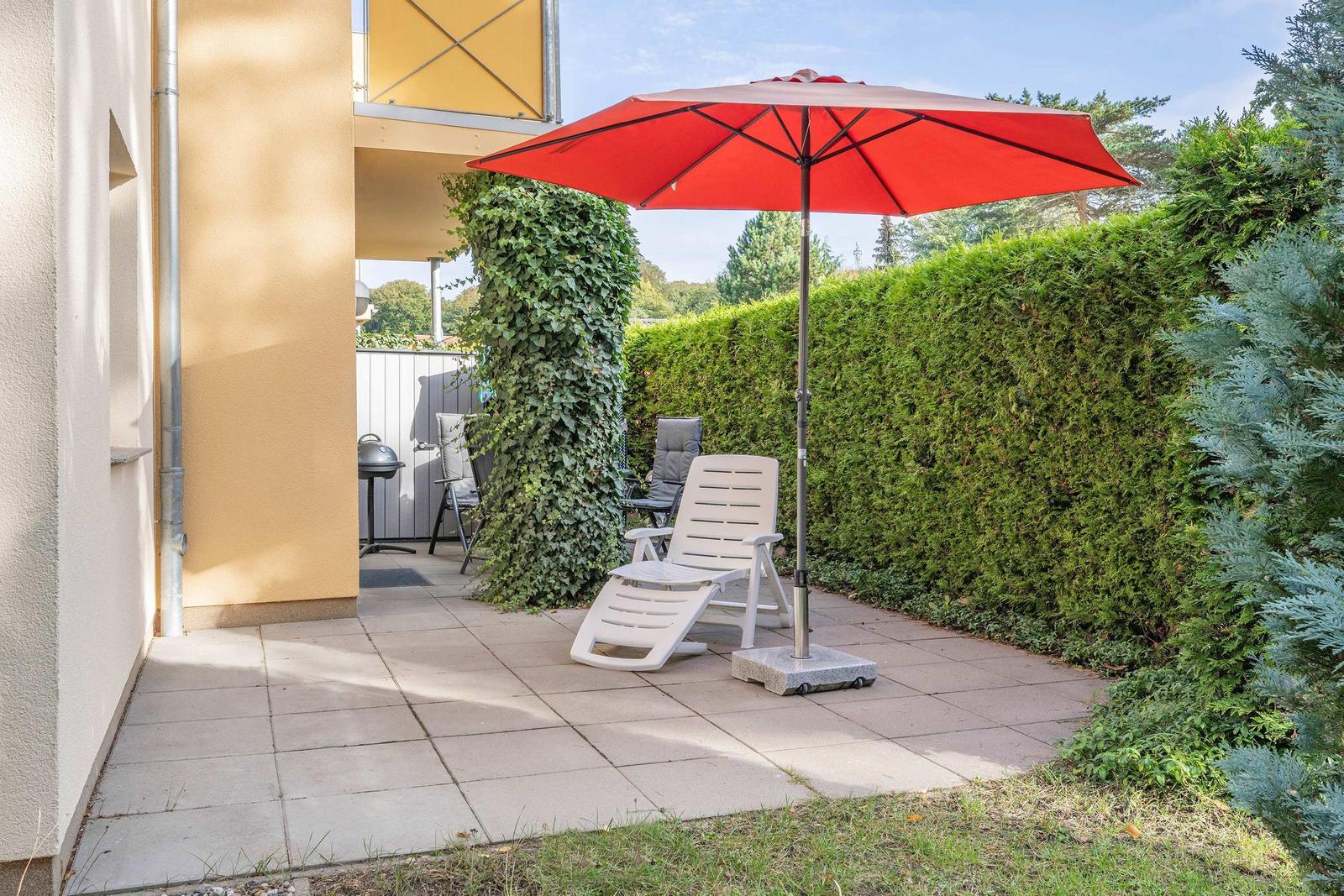 Terrace with red umbrella, lounge chair, and grill. Surrounded by green hedges.