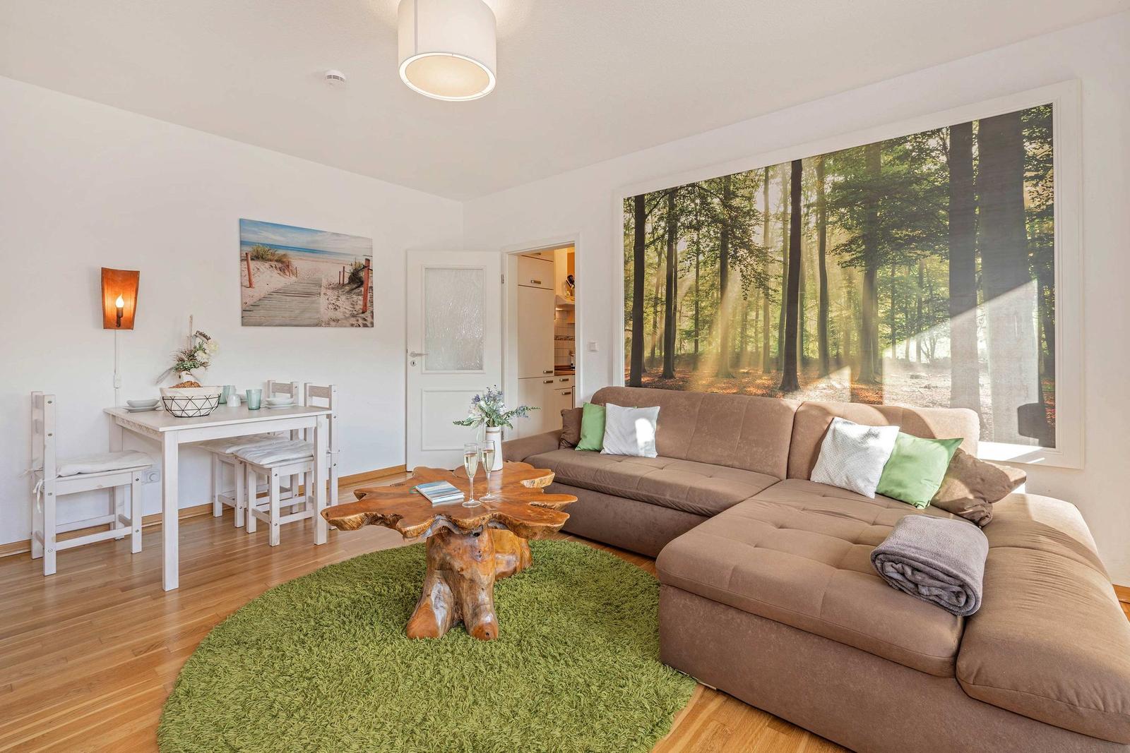 Living room with sofa, dining table, and forest wall mural. Wooden floor and green rug.