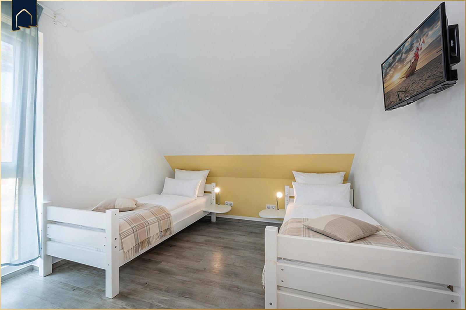 Zwei Einzelbetten mit Matratzen und Kissen, Wand-TV und Fenster mit Vorhängen.