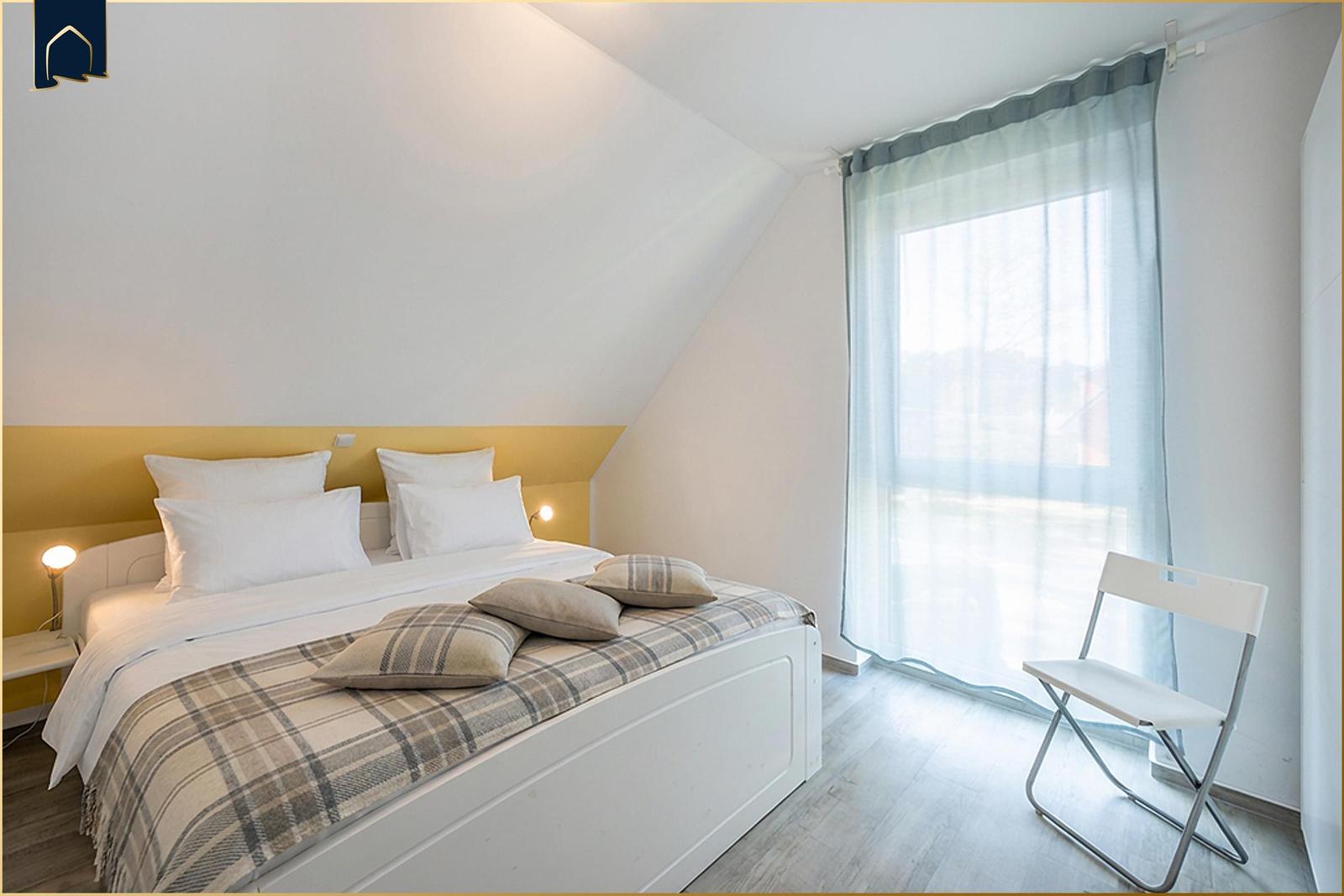 Schlafzimmer mit Doppelbett, gelbem Kopfteil und großem Fenster mit Vorhang.
