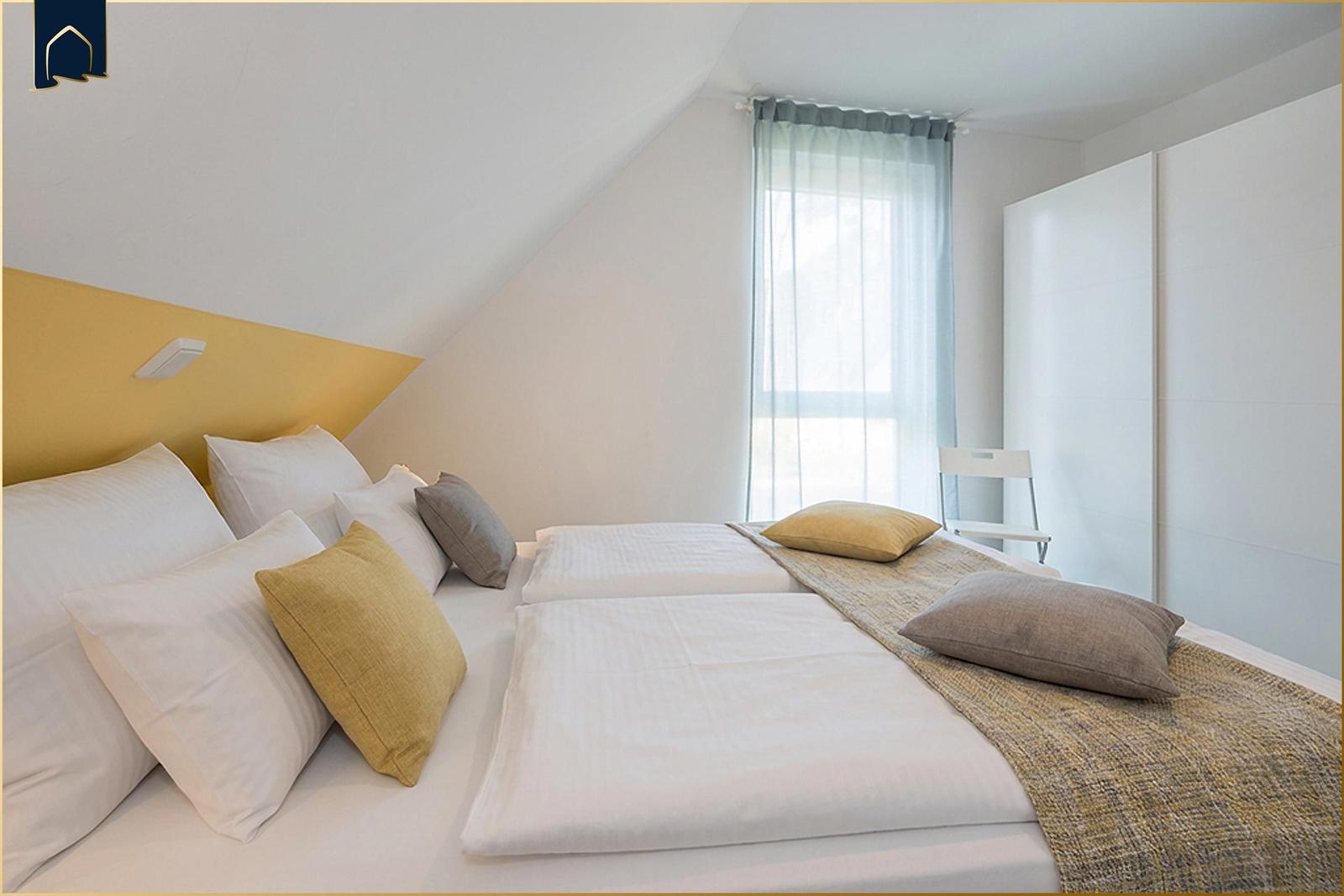 Schlafzimmer mit Doppelbett, gelbem Akzentwand und Fenster mit Vorhängen.