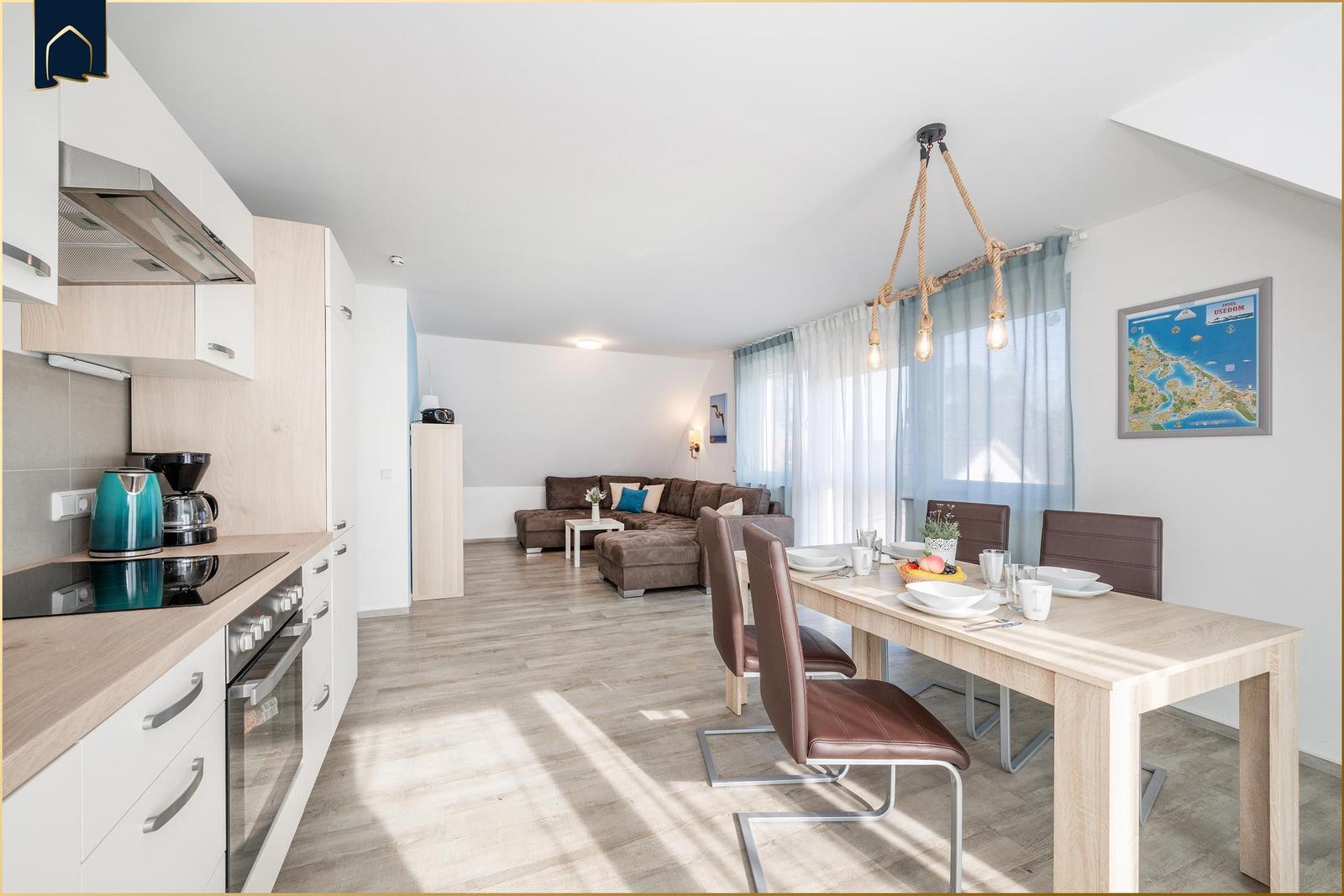 Küche, Essbereich und Wohnzimmer mit Sofa, Tisch und Stühlen. Wandbild und Vorhänge sind sichtbar.