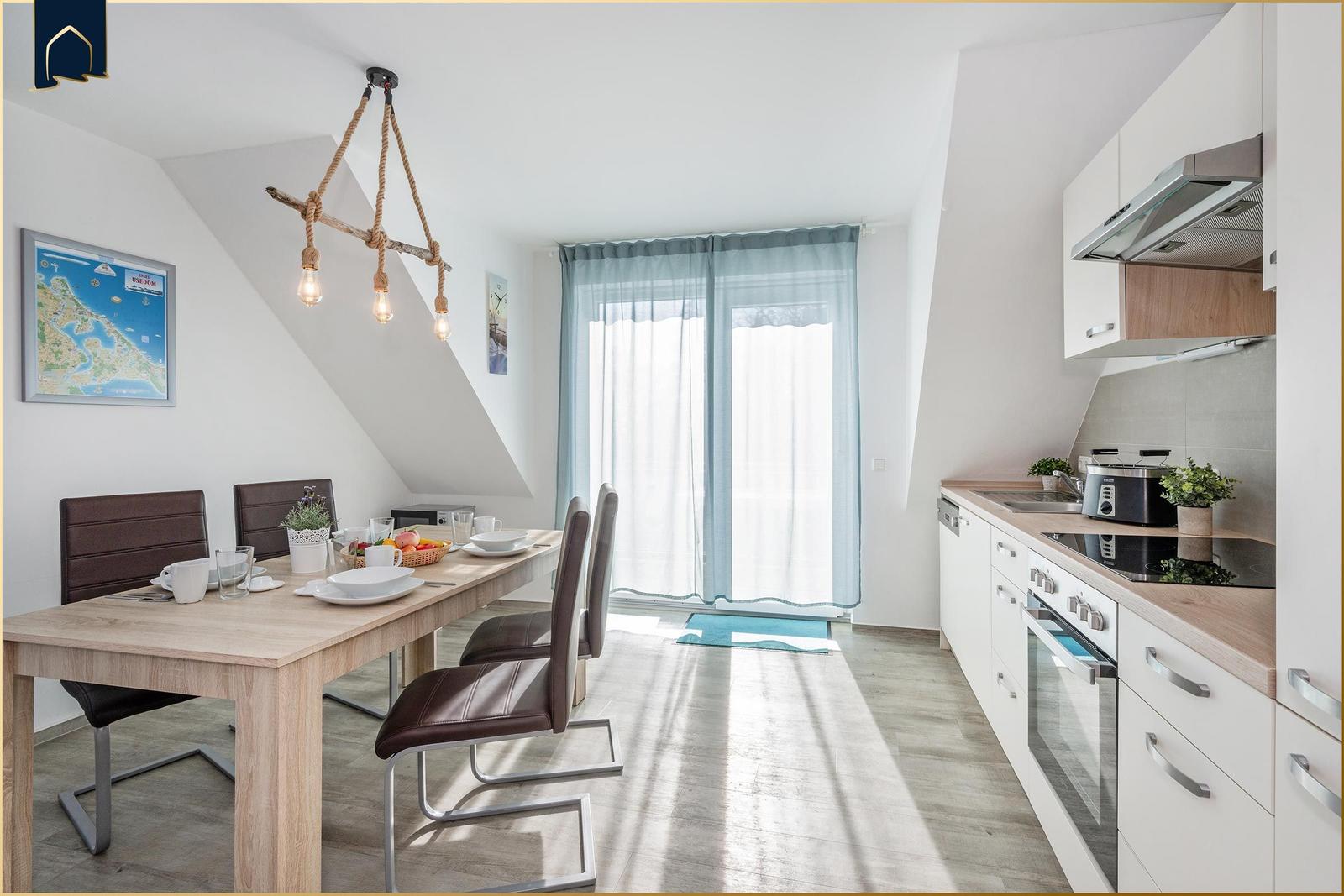 Essbereich mit Tisch, Stühlen und Küchenzeile. Fenster mit Vorhängen und Karte an der Wand.