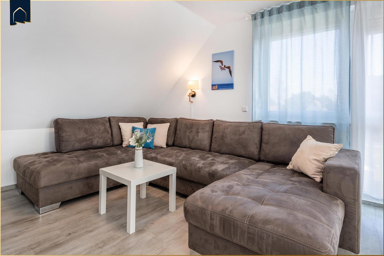Ein Wohnzimmer mit einem großen braunen Sofa, einem weißen Tisch und einem Fenster mit hellblauen Vorhängen.