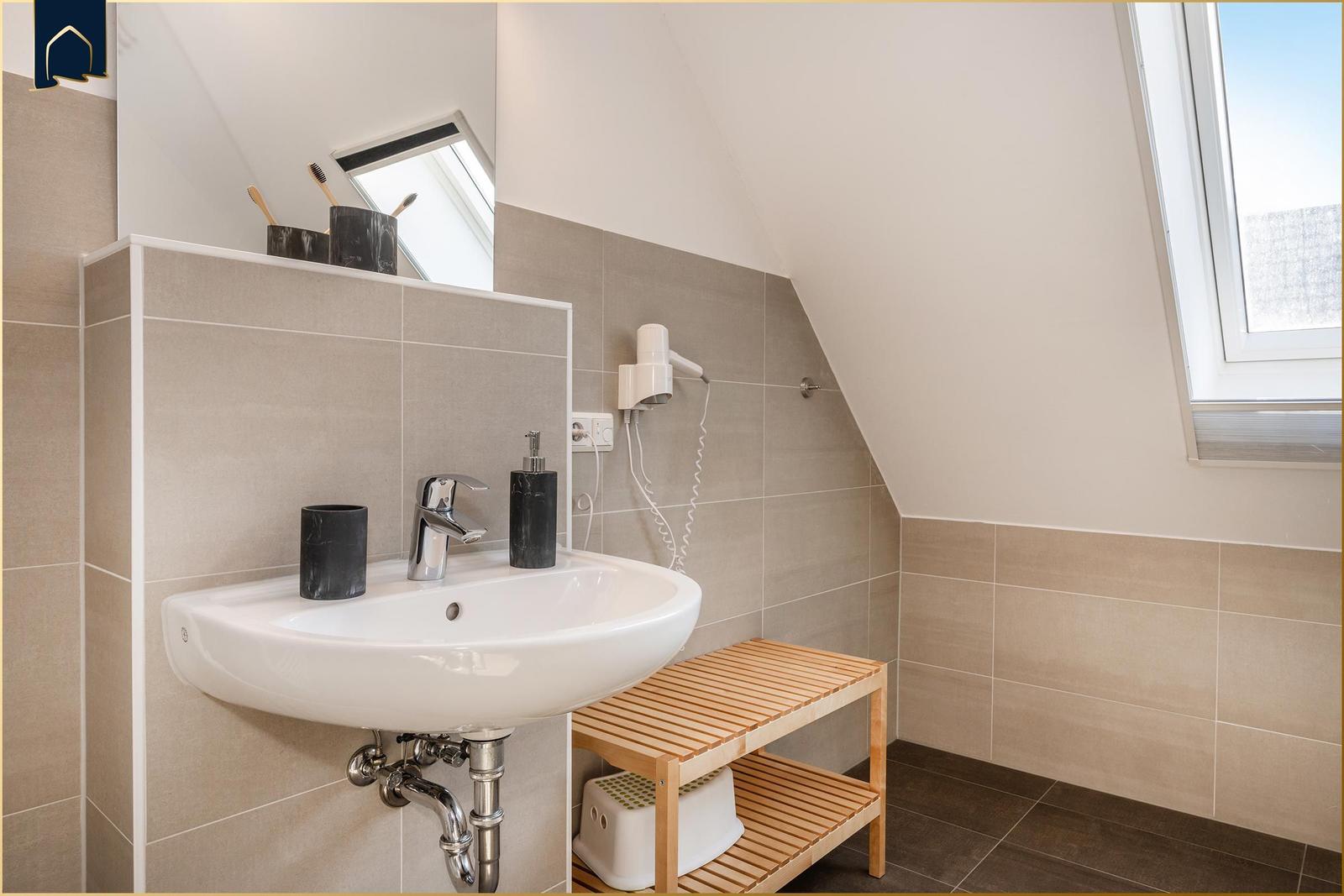 Badezimmer mit Waschbecken, Spiegel, Handtuchhalter und Holzbank unter Dachschrägen.