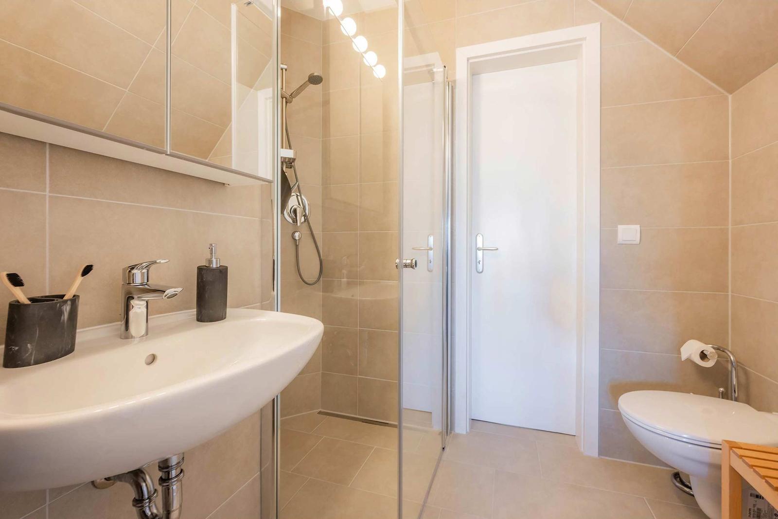 Badezimmer mit Dusche, Waschbecken und WC. Fliesenwand und Spiegelschrank.