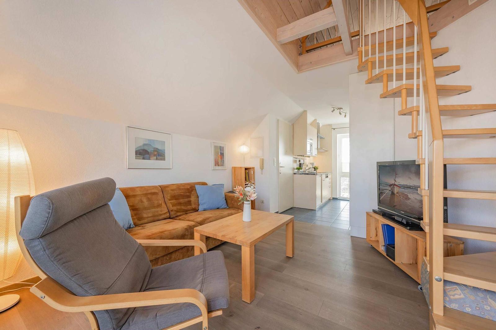 Wohnzimmer mit Sofa, Sessel, Tisch und Fernseher. Holztreppe führt zur oberen Etage.