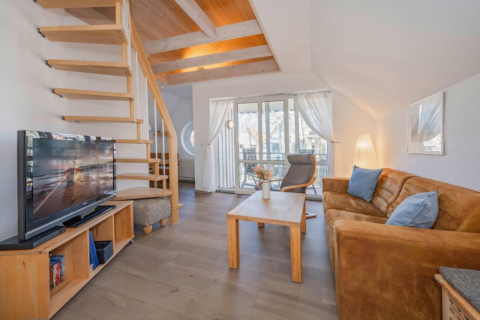 Wohnzimmer mit Sofa, Couchtisch und Fernseher. Holztreppe führt zur oberen Ebene.