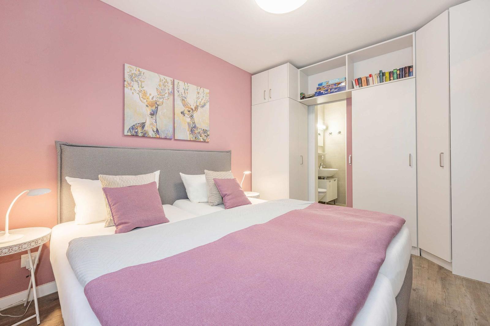 Schlafzimmer mit Bett, rosa Wand und Kleiderschrank. Badezimmer ist angebaut.