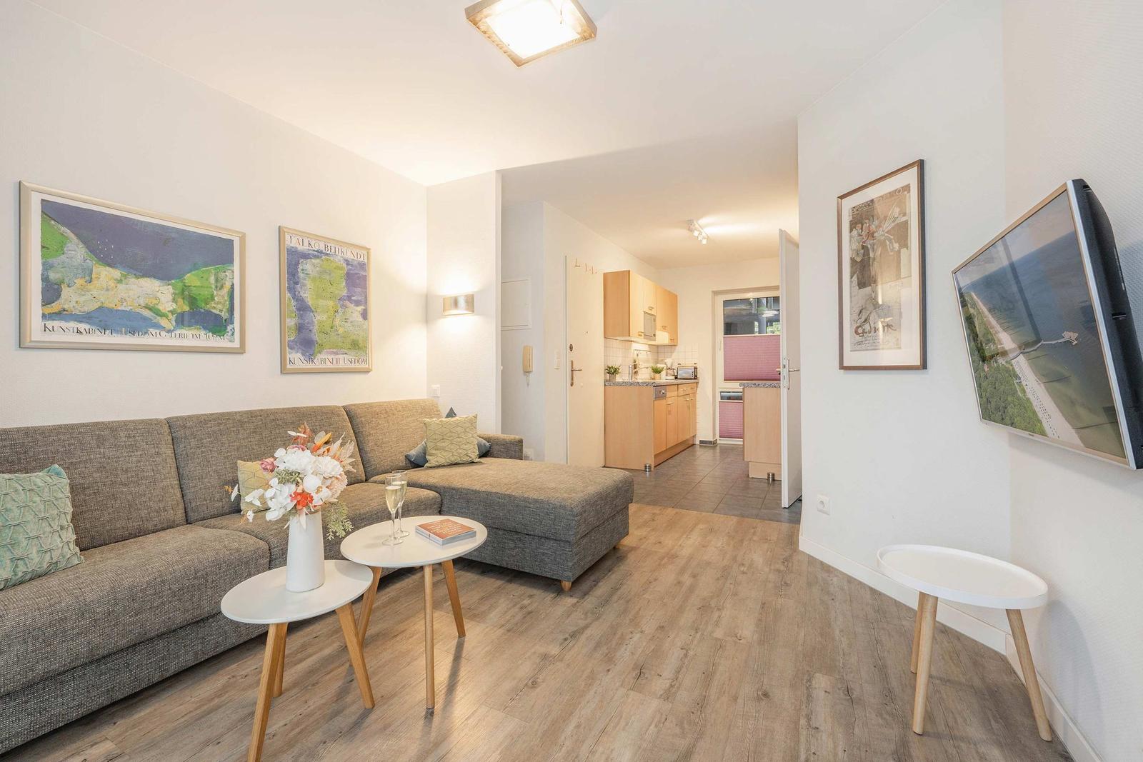 Wohnzimmer mit Sofa, Tischen und Fernseher. Küche im Hintergrund.