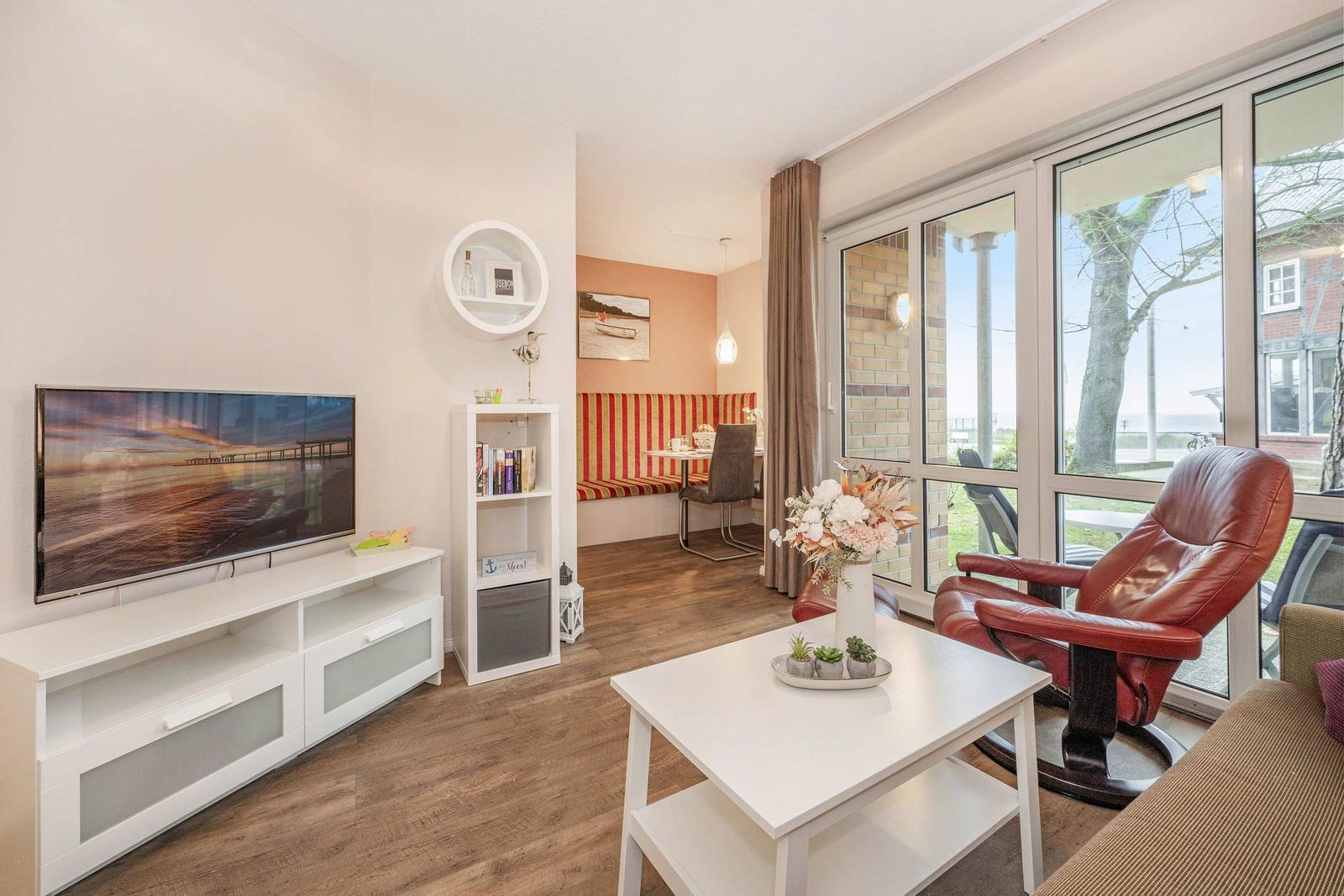 Wohnzimmer mit Fernseher, Couch und Sitzbereich. Durchgang zum Essbereich mit Sitzbank.