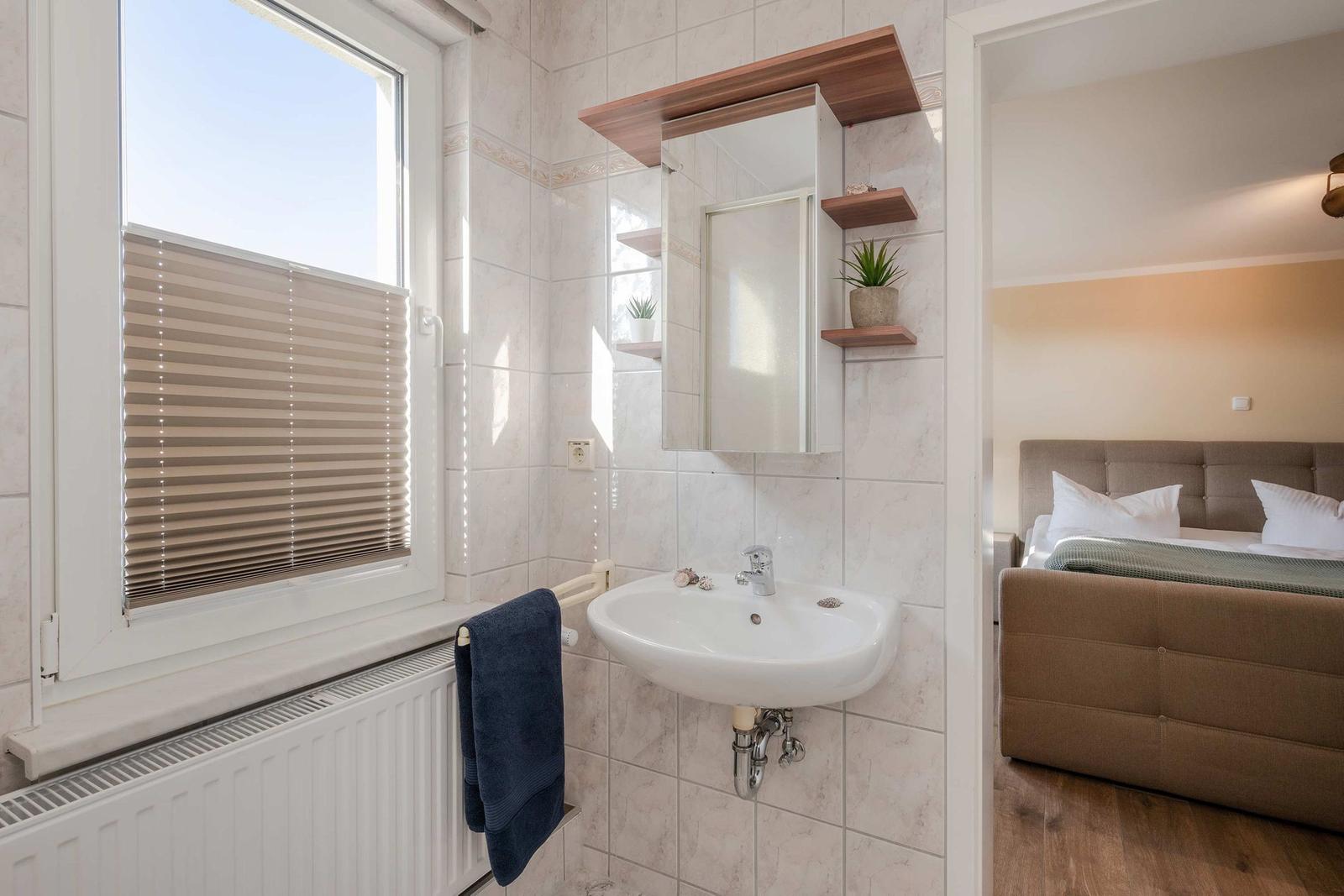 Badezimmer mit Waschbecken, Spiegel und Fenster. Im Hintergrund ist ein Schlafzimmer mit Bett sichtbar.
