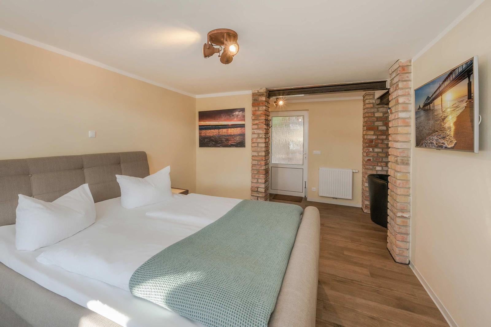 Schlafzimmer mit Doppelbett, Holzboden und Wandbildern. Zentraler Durchgang mit Ziegelwänden.