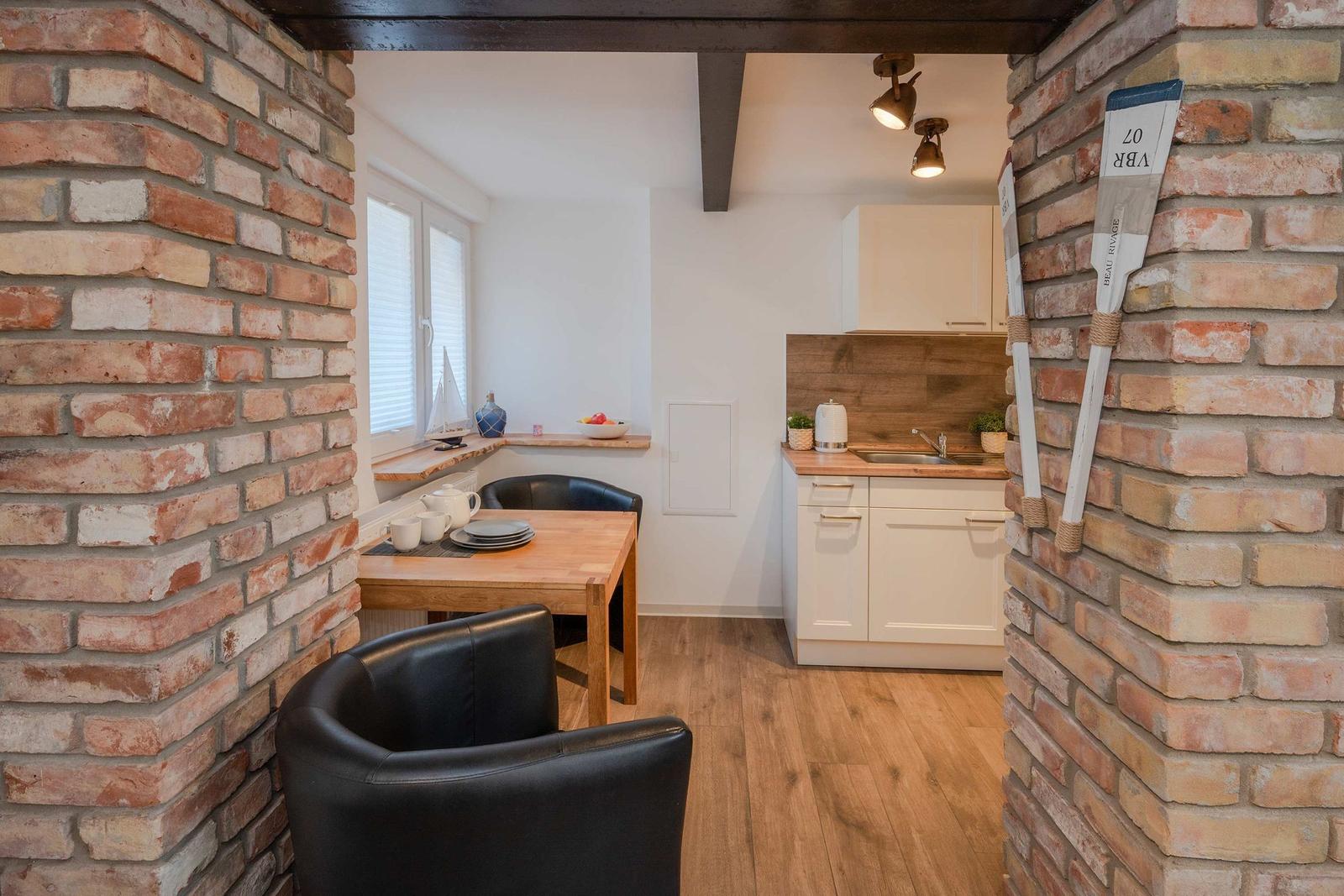 Küche mit Esstisch, Holzboden und Backsteinwänden. Fenster mit Jalousie und kleine Pflanzen.