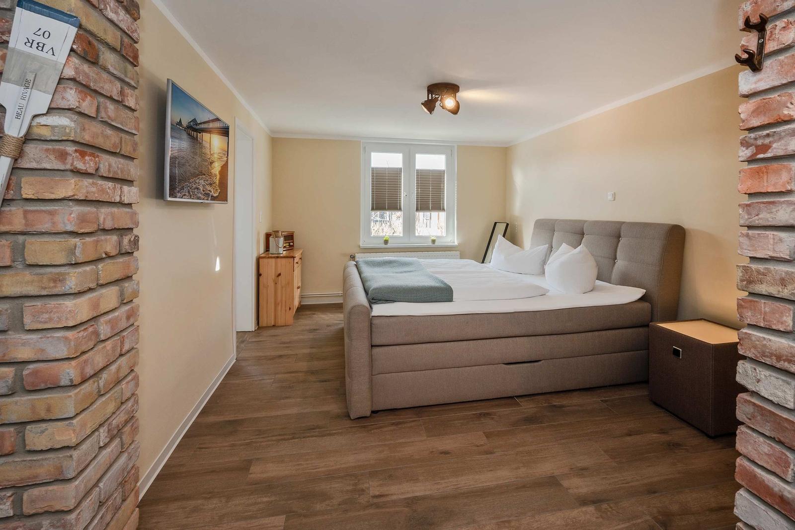 Schlafzimmer mit Bett, Holzboden und Fenster. Zwei Steinwände flankieren den Raum.