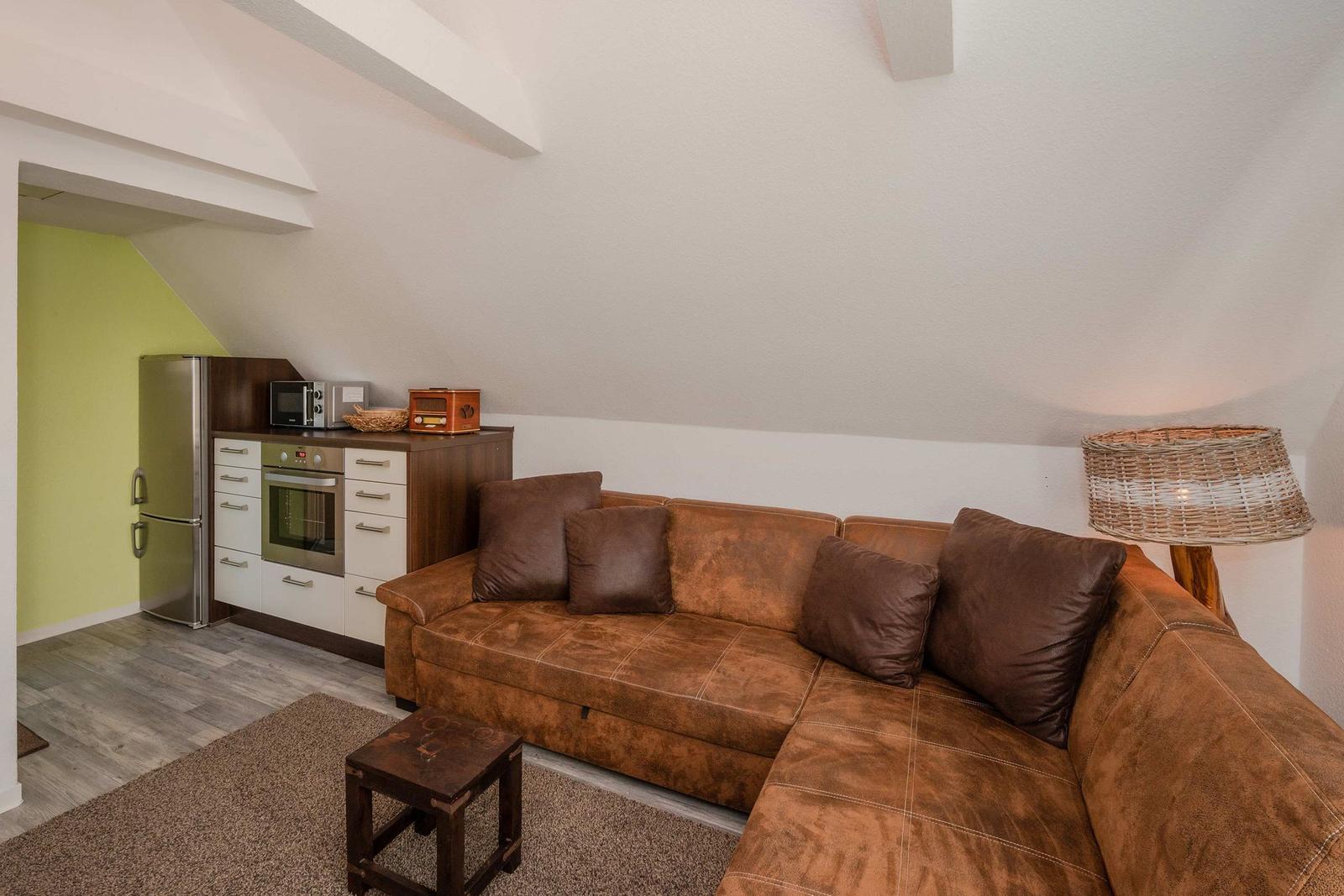 Wohnbereich mit braunem Sofa, Küchenzeile und Stehlampe unter schrägen Wänden.