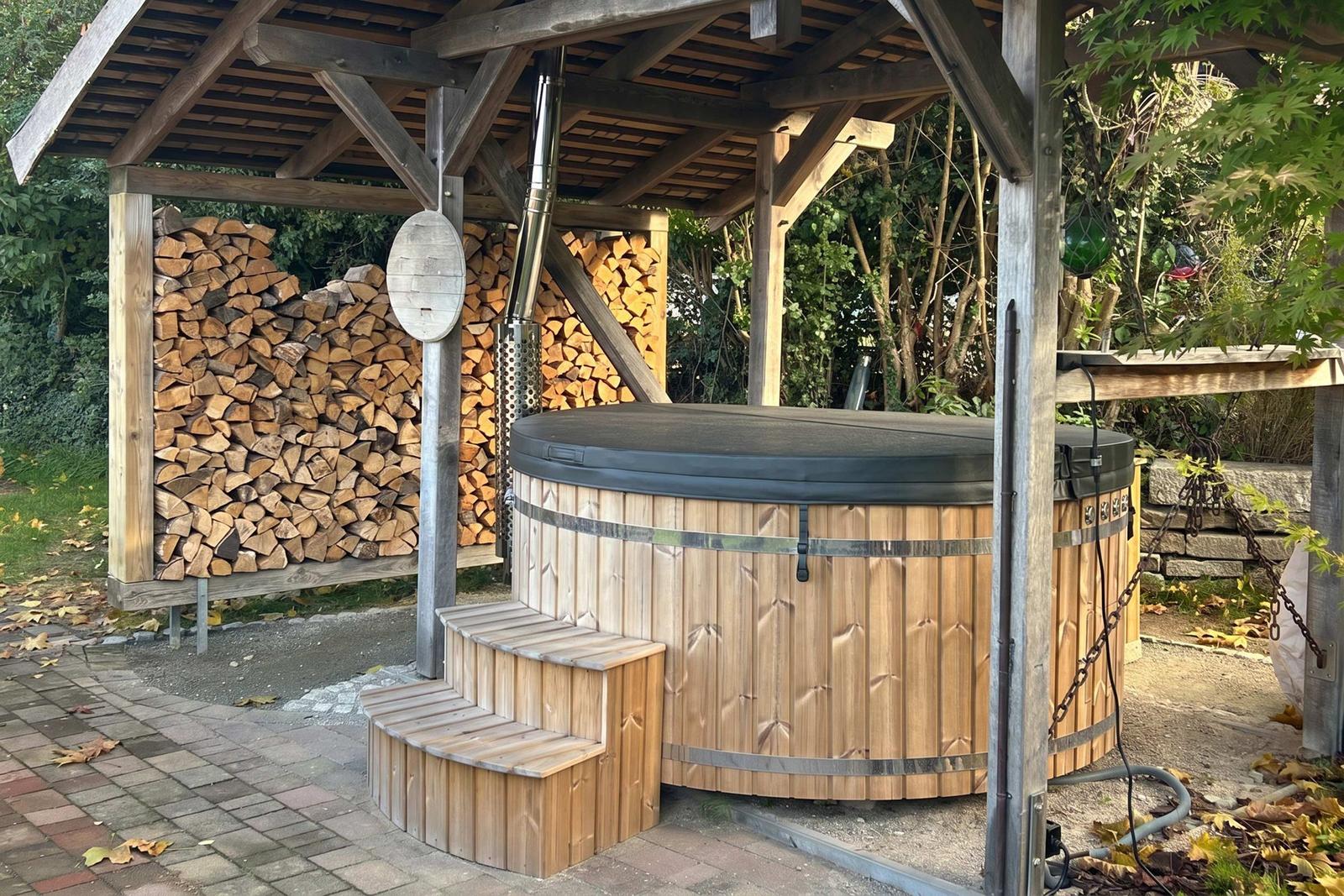 Holzüberdachter Whirlpool mit Sitzstufen und Holzstapel im Hintergrund.