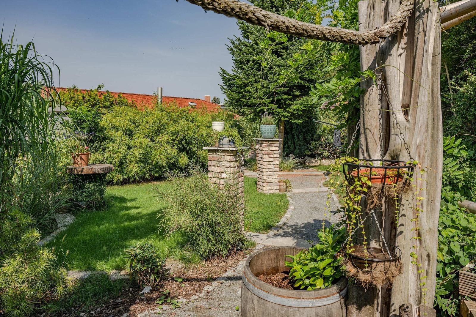 Garten mit Pflanzen, Steingartenweg und Holzpfosten. Blick auf das Haus mit rotem Dach.