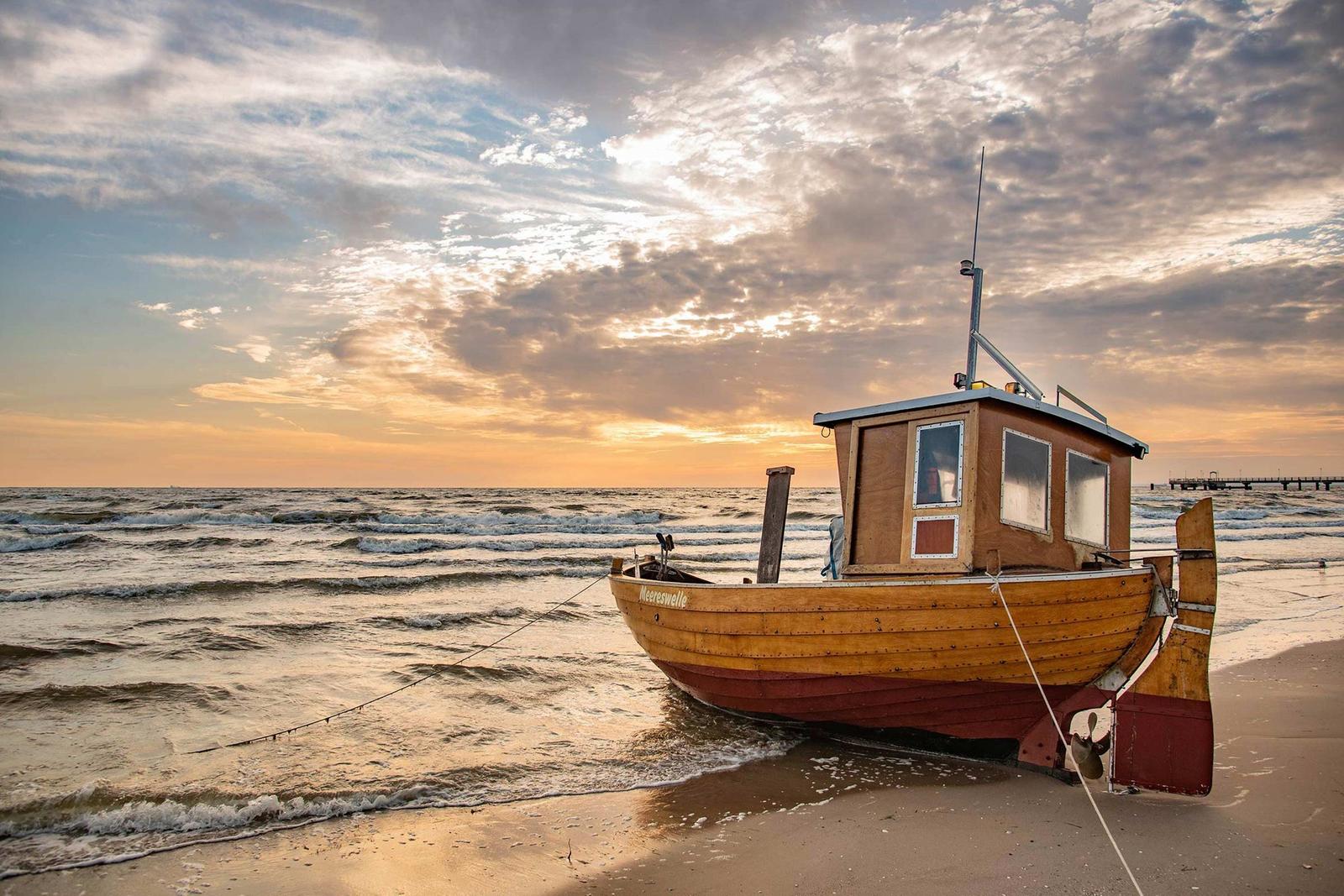 Ein hölzernes Boot liegt am Strand, im Hintergrund See und Himmel bei Sonnenuntergang.