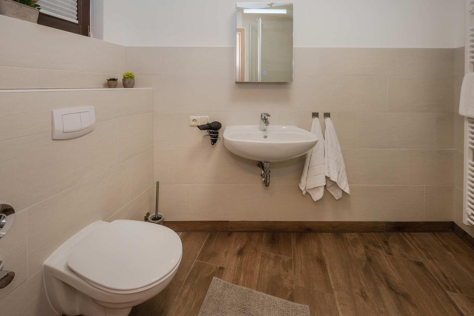 Badezimmer mit Toilette, Waschbecken und Spiegel. Holzfußboden und weiße Fliesen.