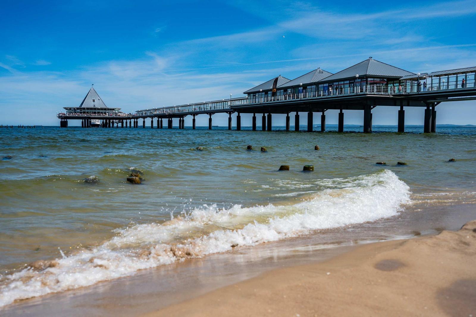 Strand mit Wellen, Steine im Wasser und Pier mit Dächern und Besuchern.